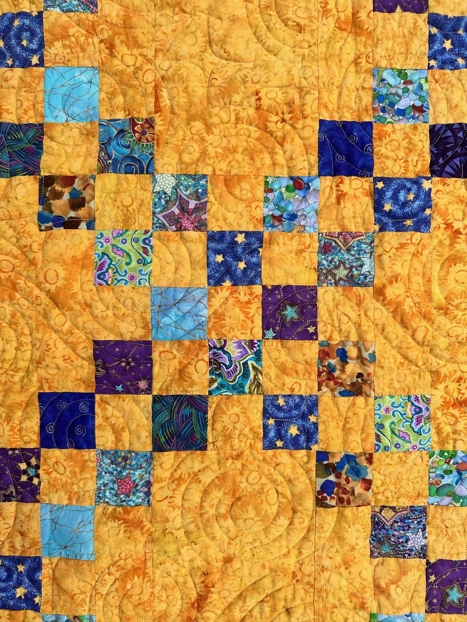Close-up view of a colorful quilt with a patchwork design, featuring yellow, blue, purple, and multicolored fabric squares with various patterns and intricate stitching.
