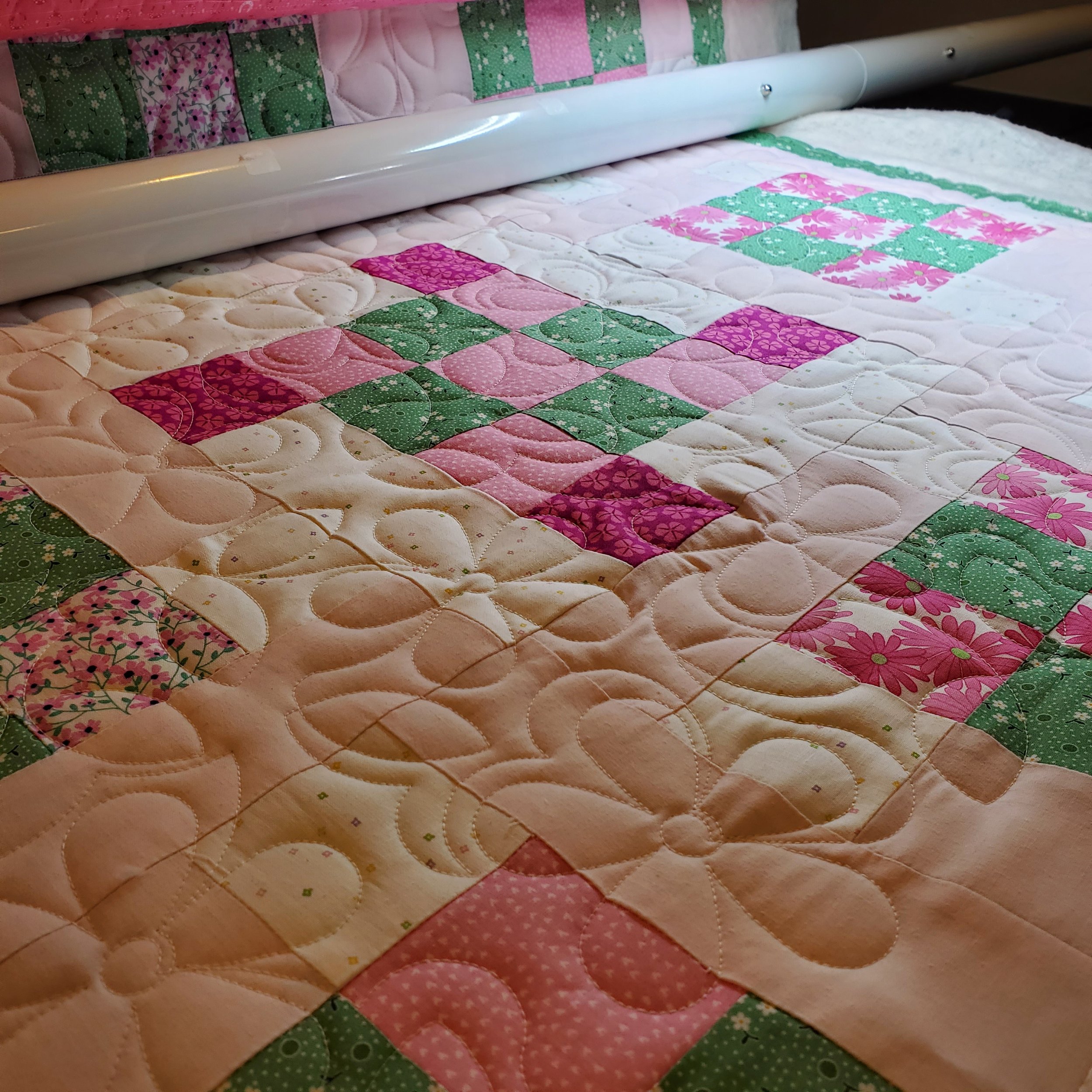 Close-up of a colorful quilt with pink, green, and white patterns, featuring floral and geometric designs, quilted with swirling and straight stitches.