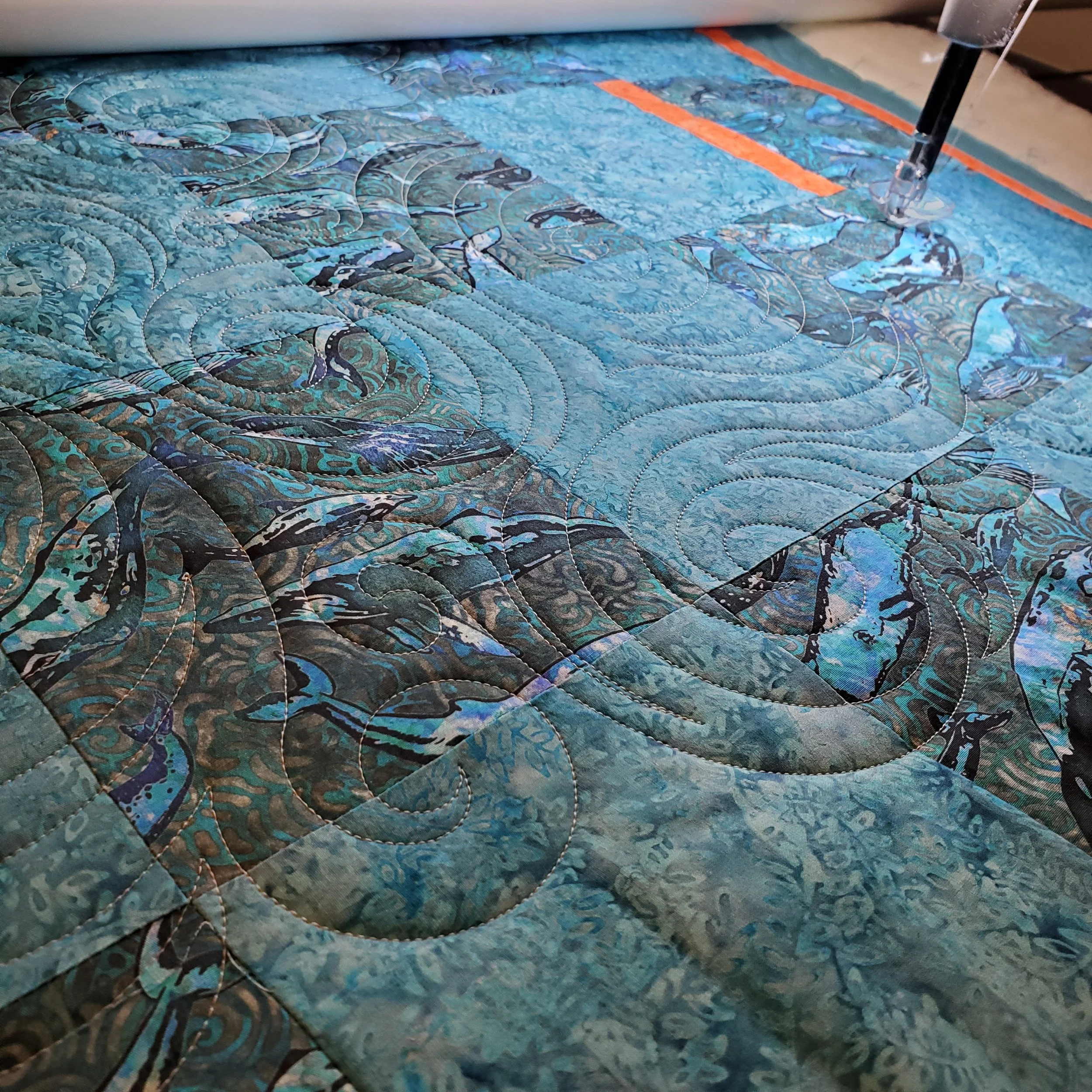 Close-up of a detailed blue and black quilt with swirl and wave patterns, under a sewing machine needle.