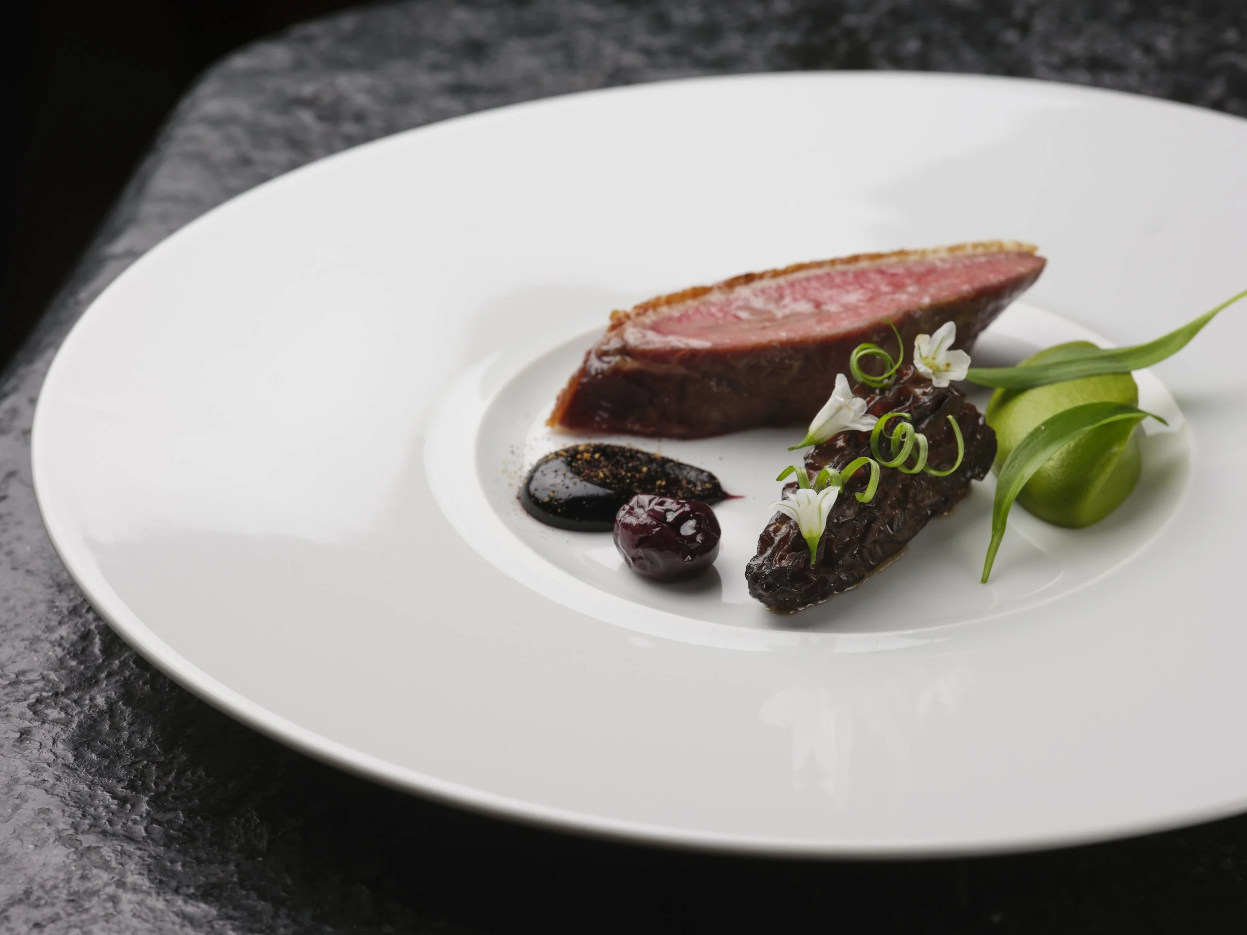 A gourmet dish featuring a slice of medium-rare steak with a dark sauce, garnished with green onions, edible flowers, and a side of vegetables on a white plate.