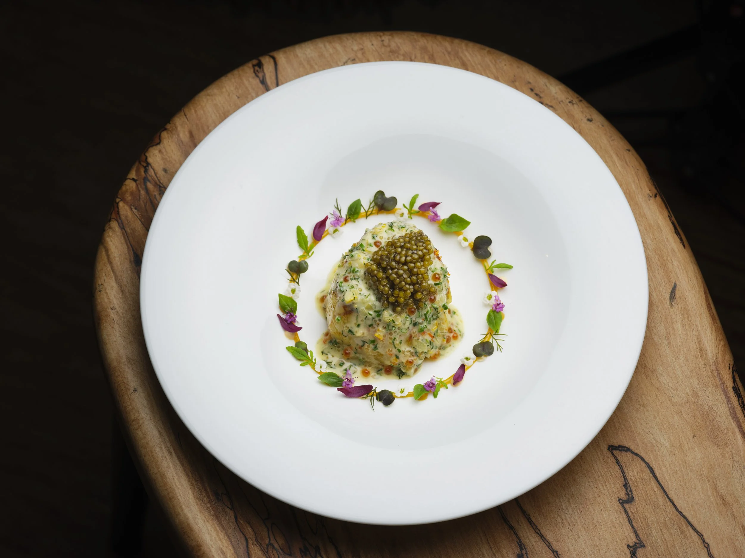 A plated gourmet dish consisting of a bbq scallop topped with small green caviar, garnished with herbs, flowers, and small dark spheres, served on a white plate with a wooden base.