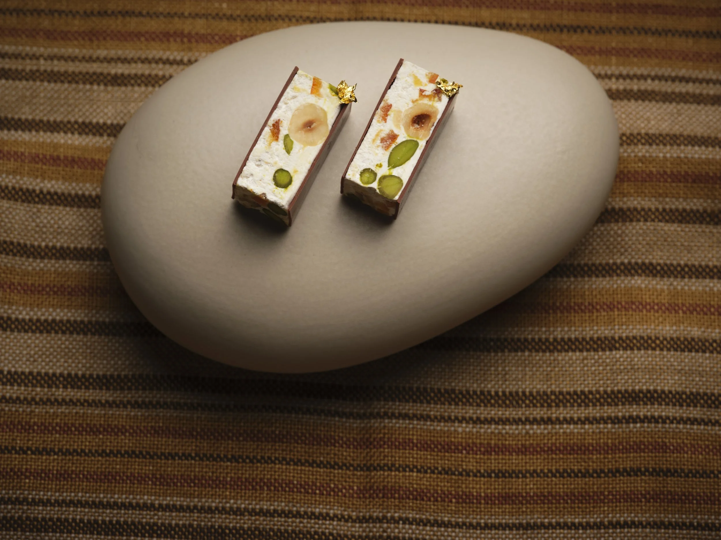 Two rectangular pieces of decorative nougat with white, orange, and green embedded candies, topped with gold leaf, are placed on a smooth, white, oval-shaped stone on a brown striped fabric surface.