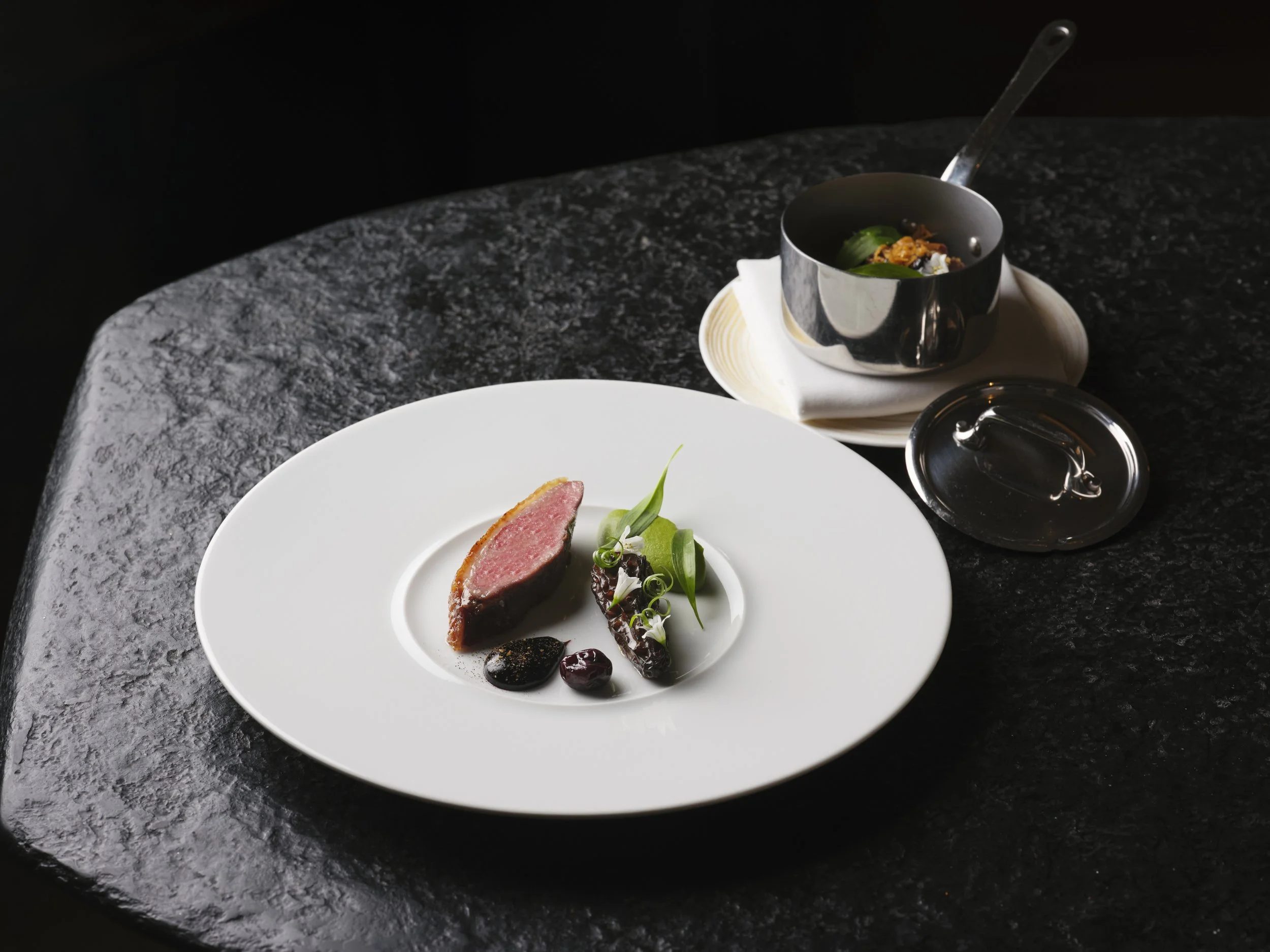 A gourmet dish featuring a cut of  with a pink interior, garnished with green herbs and accompanied by a dark sauce and small dollops of a red sauce, served on a white plate. In the background, there is a small black pot with a lid, placed on a white