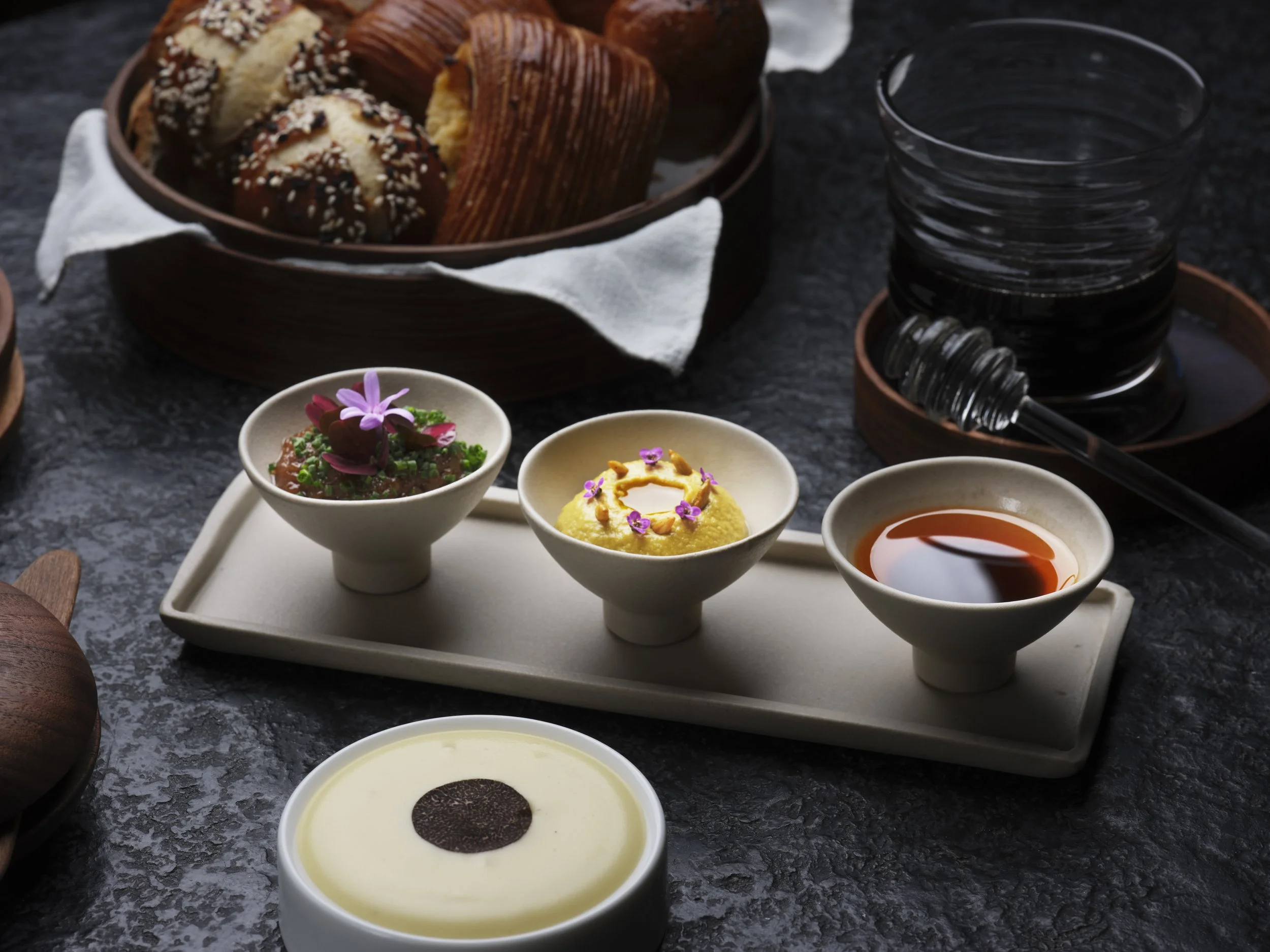 A dessert platter with various small bowls containing different sweet dishes, including custard topped with a cookie, a green garnish with flowers, and a yellow pudding with flower petals, alongside slices of bread and a glass of water on a dark marb