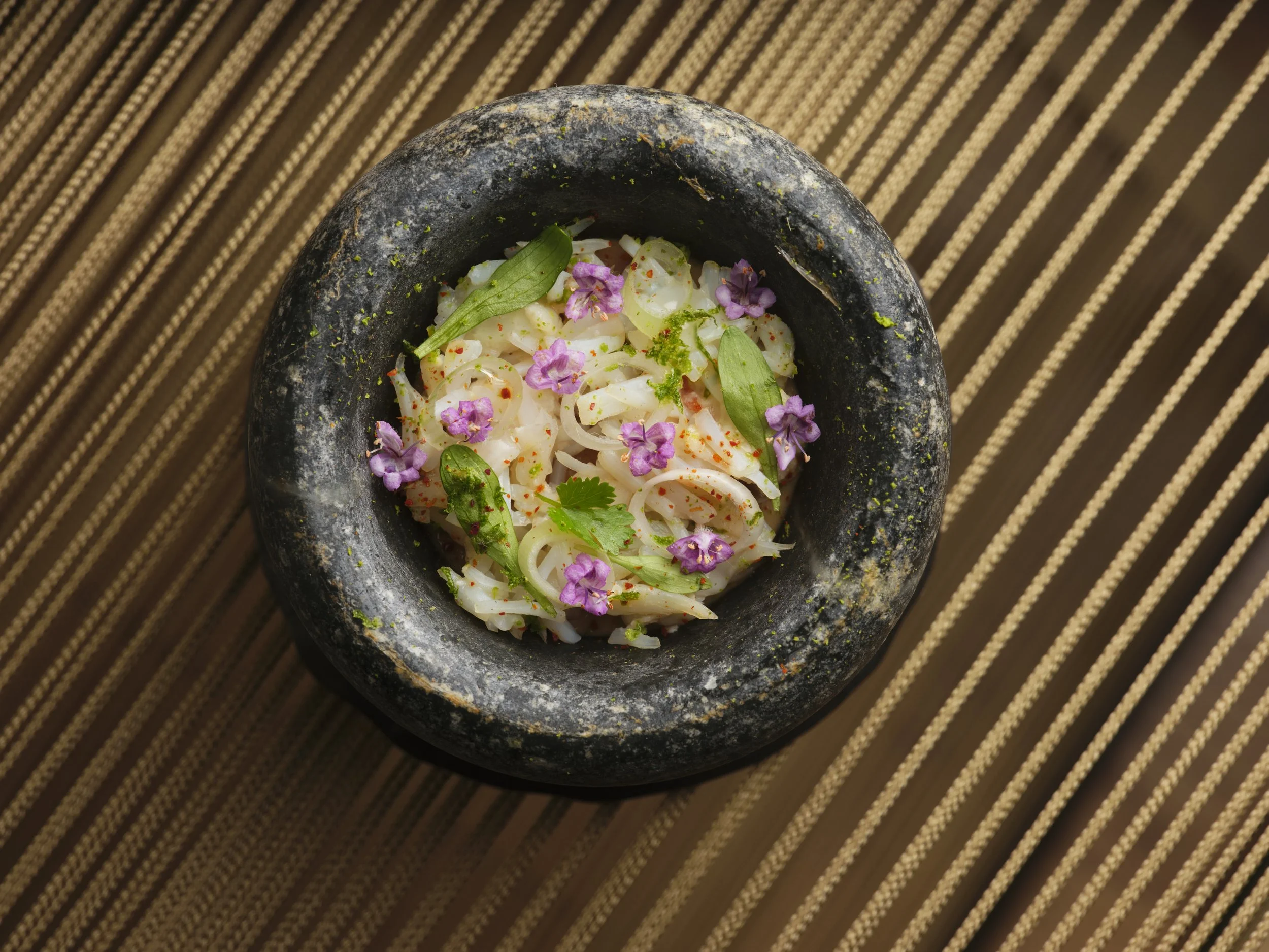 A close-up of a traditional Mexican molcajete filled with a cooked onion salad garnished with small purple and green herbs, placed on a wooden striped surface.