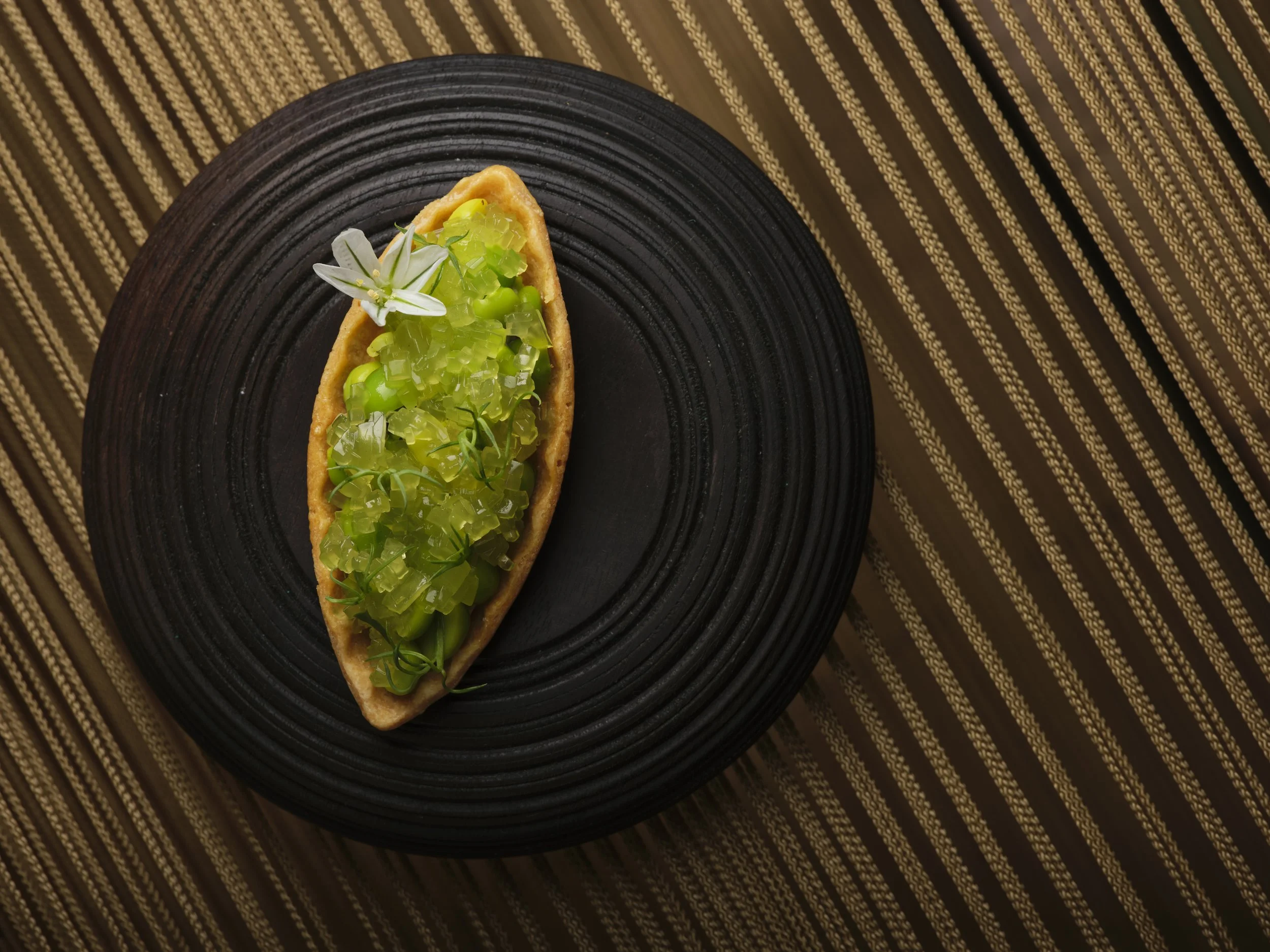 A pastry boat filled with green jelly-like dessert topped with edible flowers and herbs, served on a black textured plate and a striped brown tablecloth.