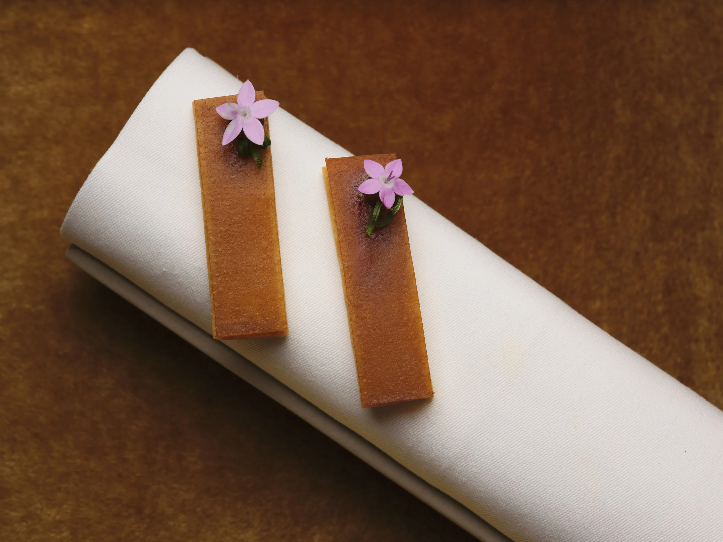 A white cloth napkin is folded and adorned with two narrow brown paper strips, each topped with a small pink flower with five petals and green leaves, on a brown textured surface.