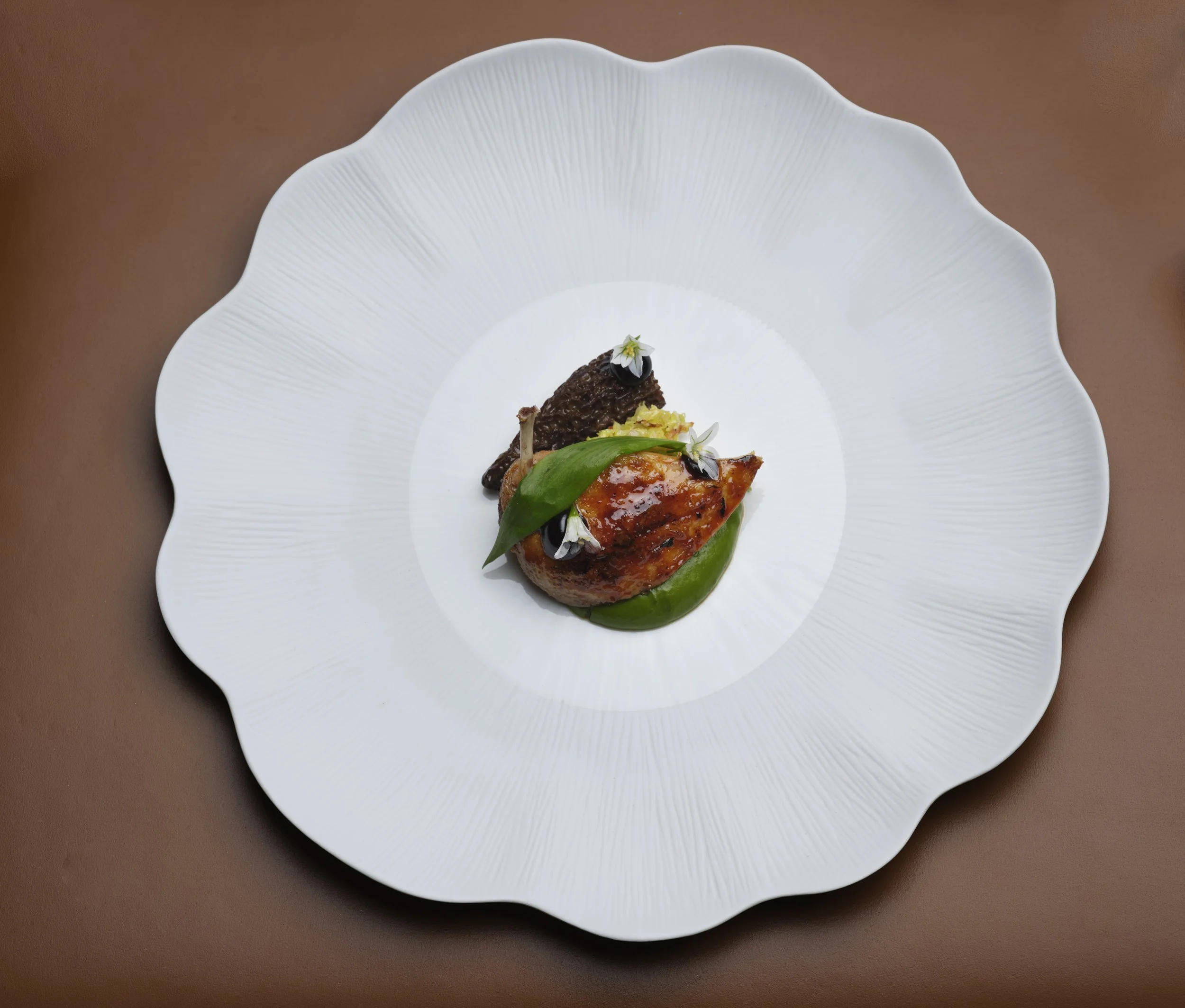 A white decorative plate with uneven edges, containing a poached roasted quail with morrell mushrooms garnished with edible flowers, herbs, and sauce.