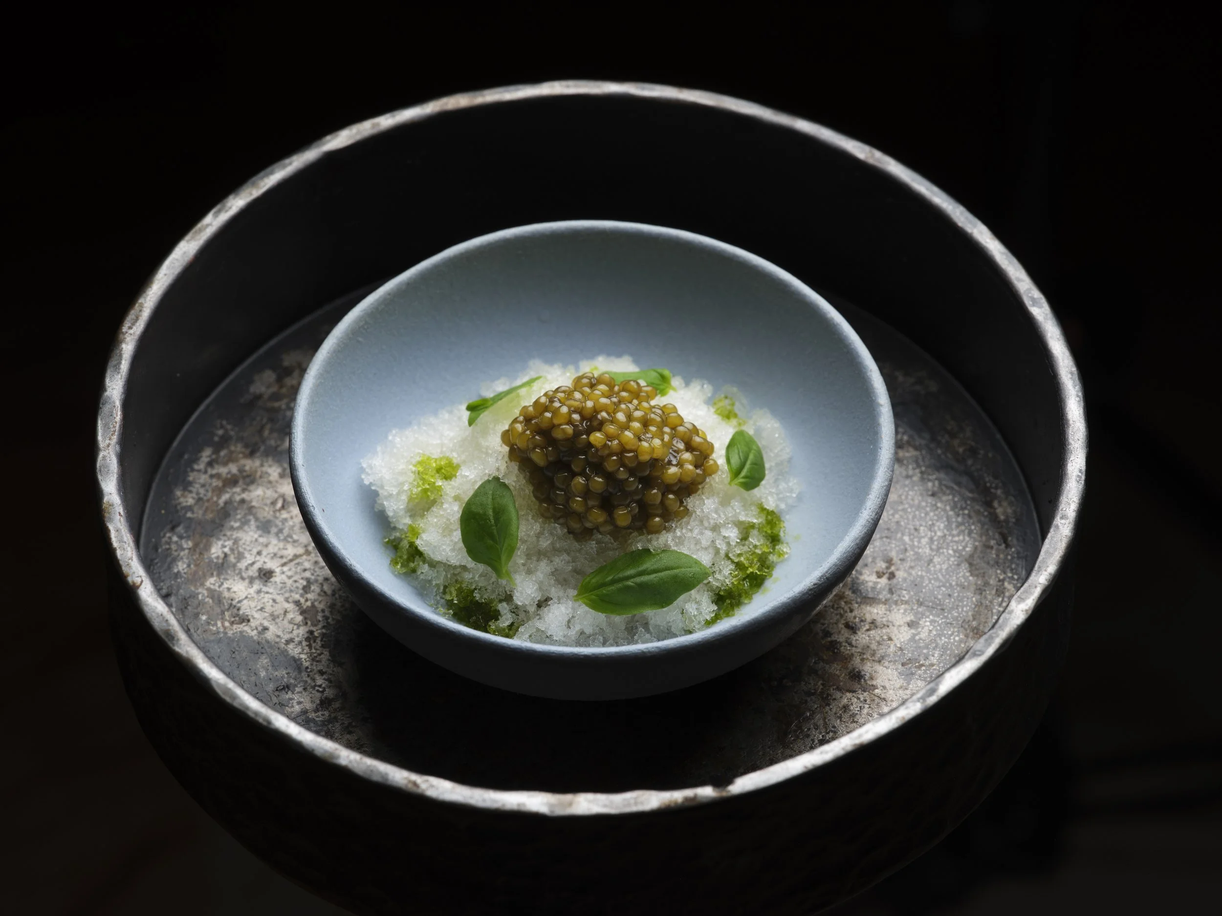 A dish with caviar, garnished with green herbs, served in a gray bowl on a dark textured surface and a black background.