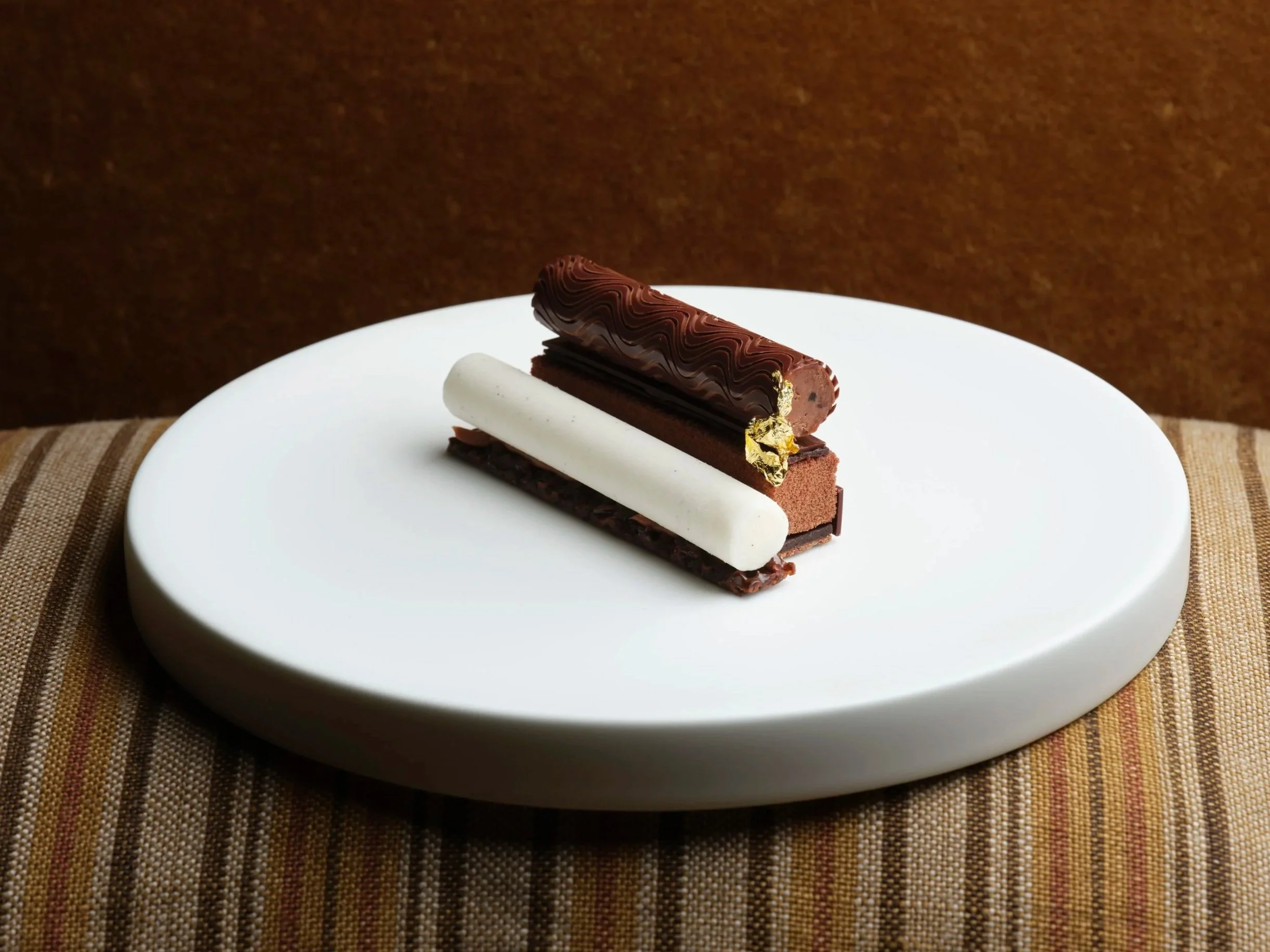 A chocolate pastry with a decorative dark chocolate glaze and gold leaf garnish, a white cylindrical pastry, and a rectangular chocolate base, all presented on a white plate.