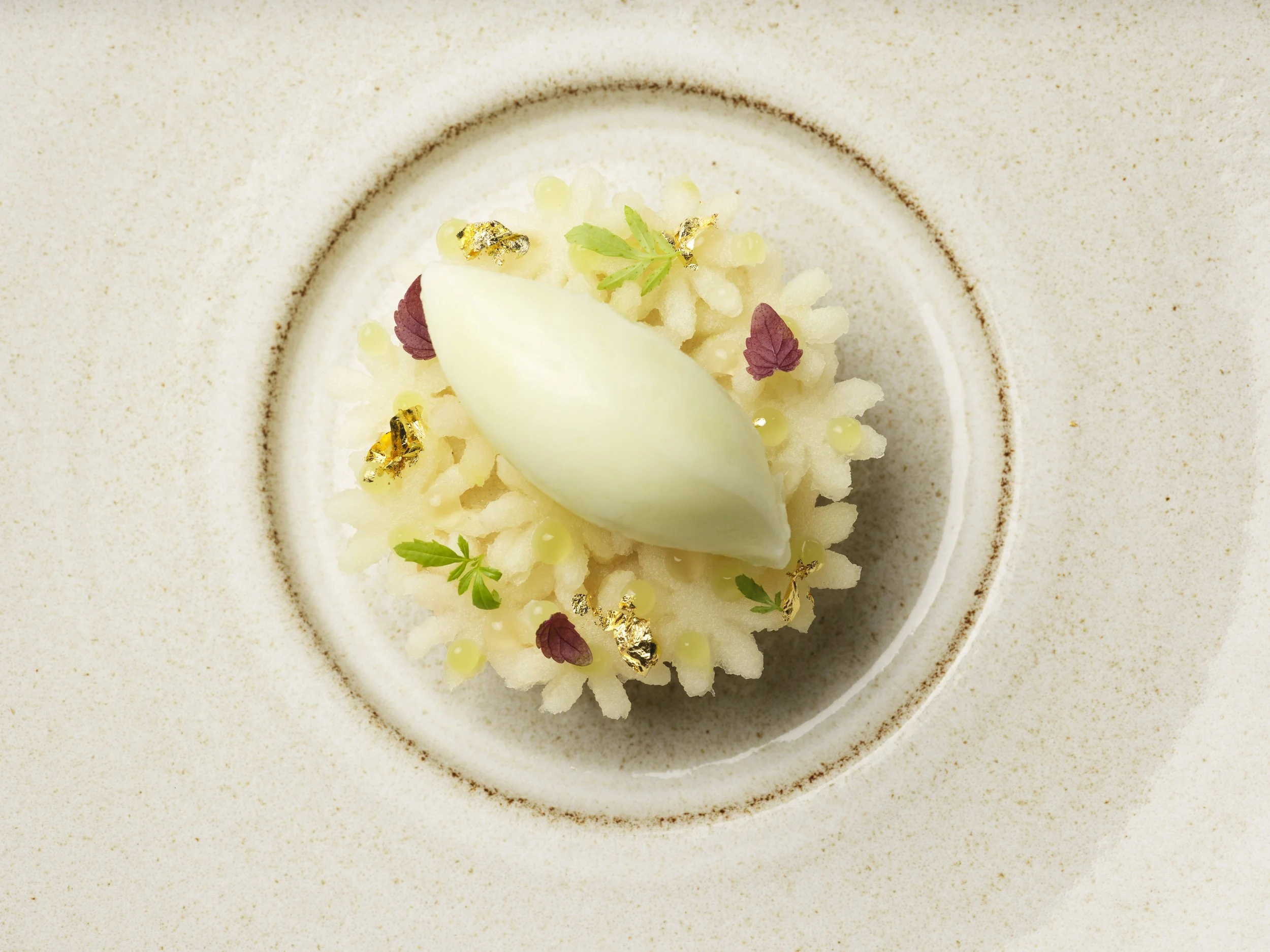 A dessert featuring white chocolate mousse, edible gold flakes, small green leaves, and a white crispy wafer, arranged on a white plate with a slightly speckled rim.