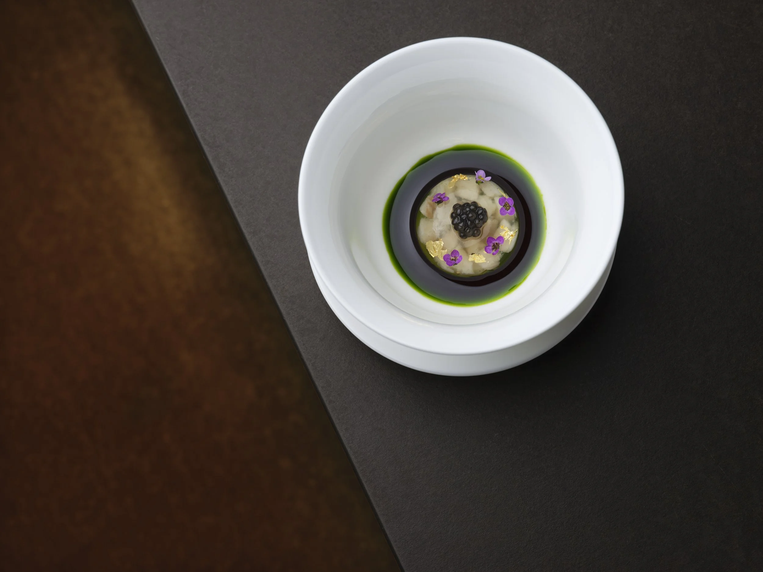 A white bowl with a colorful food presentation, including small purple flowers, black caviar, and edible gold flakes in a dark sauce, on a black and wood surface.