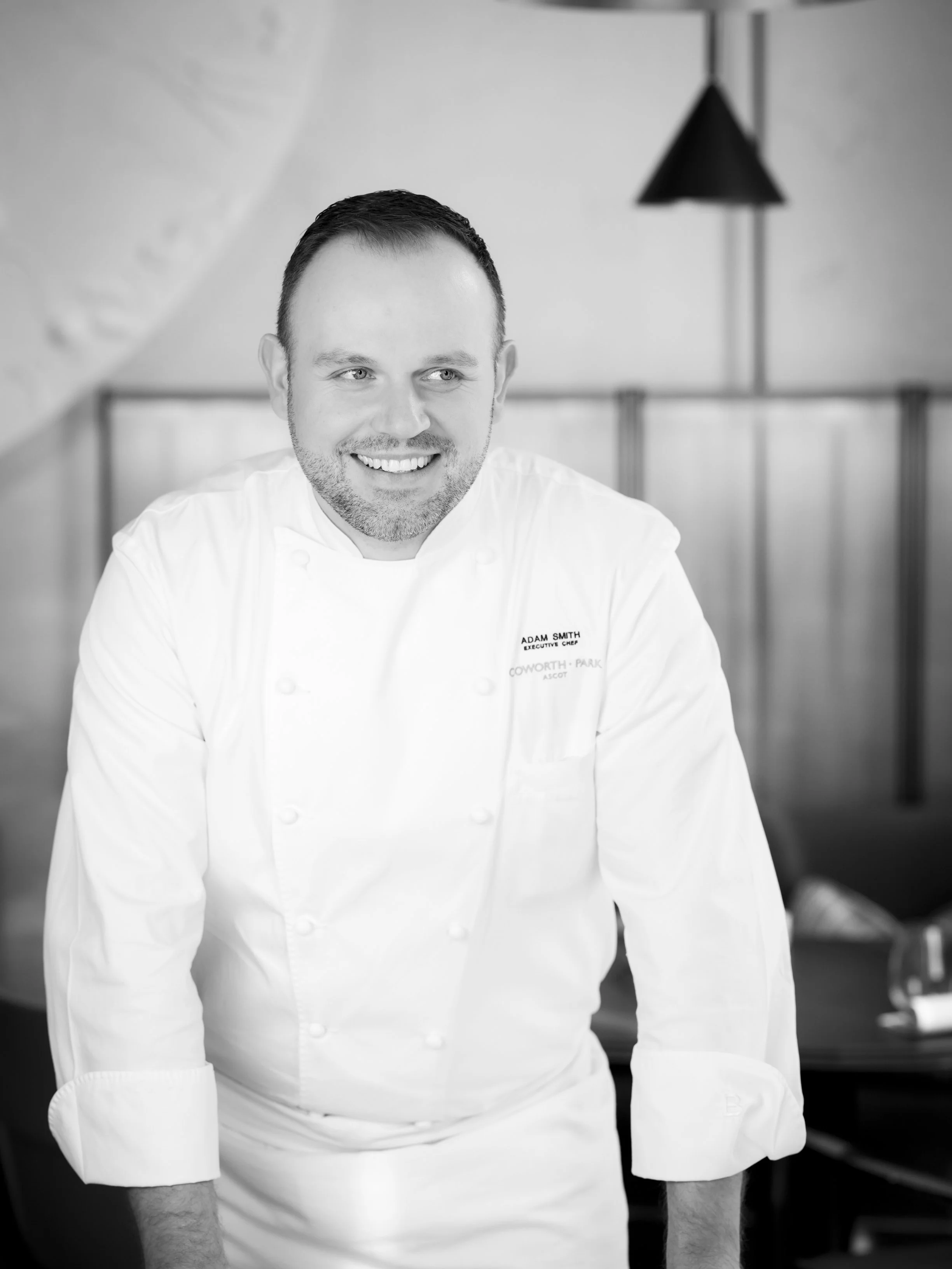 Black and white photo of a smiling chef in a restaurant kitchen. He is wearing a white chef's coat with his name and title embroidered on it, and has short dark hair and a beard. The background shows kitchen equipment.