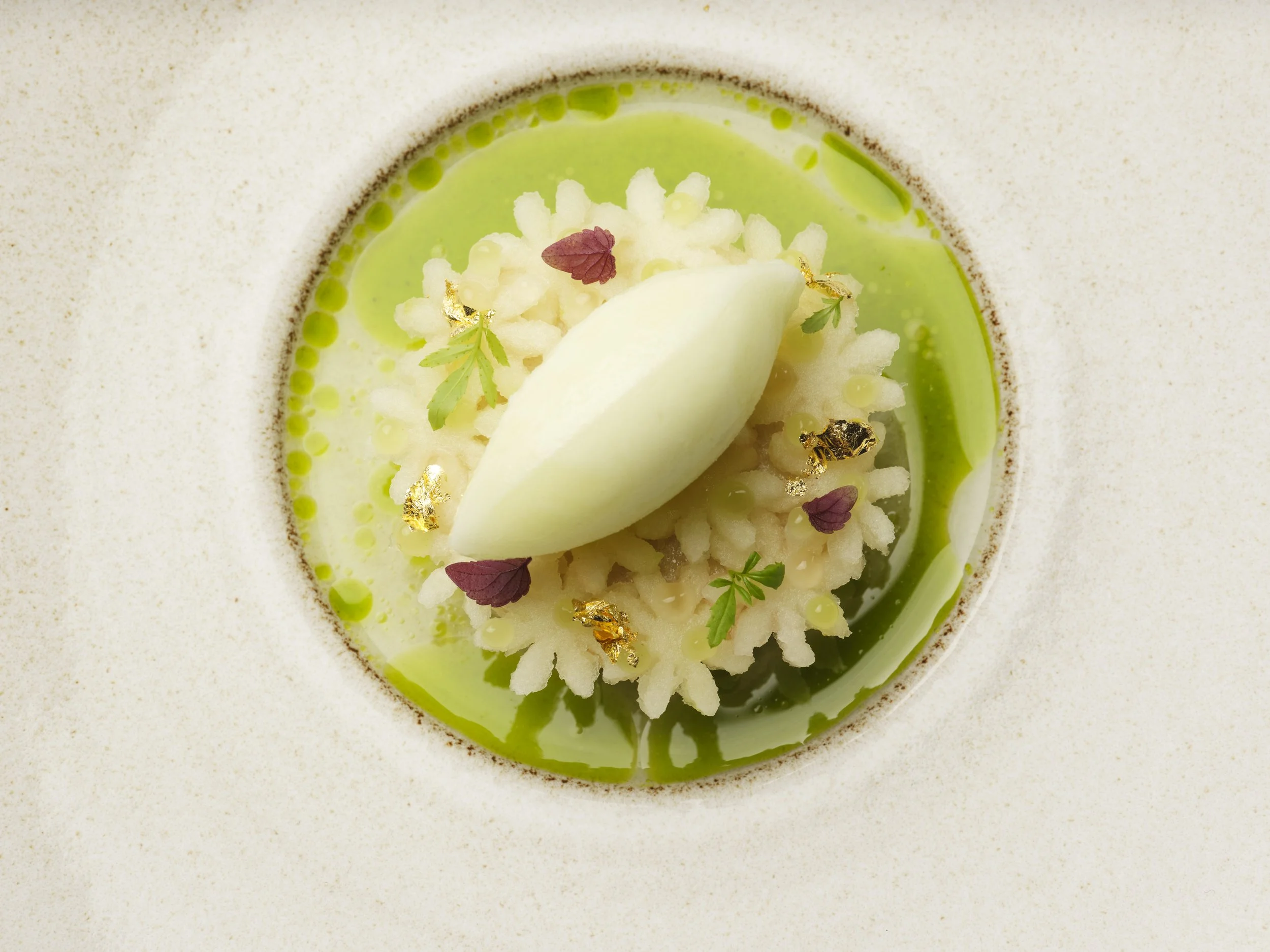 A close-up of apple plated dessert featuring green sauce, white chocolate, edible gold leaf, microgreens, and colorful edible flowers.