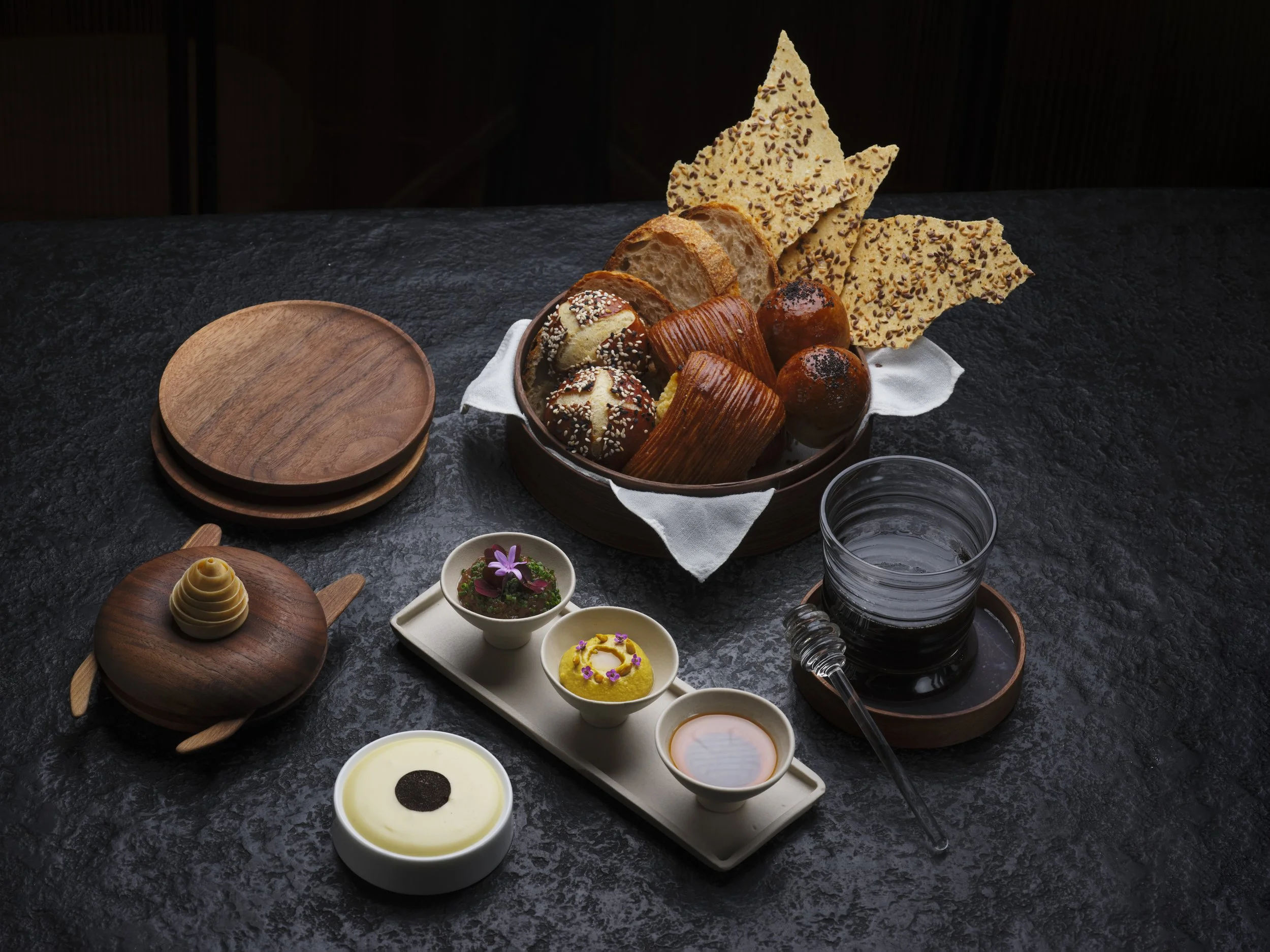 A dark textured table features a wooden platter with a small dessert topped with a flower, a bowl of yellow custard with a chocolate disc, a white rectangular tray with four small cups containing different sauces or creams, a glass of water, and a round bowl holding bread, cookies, and decorative crackers.