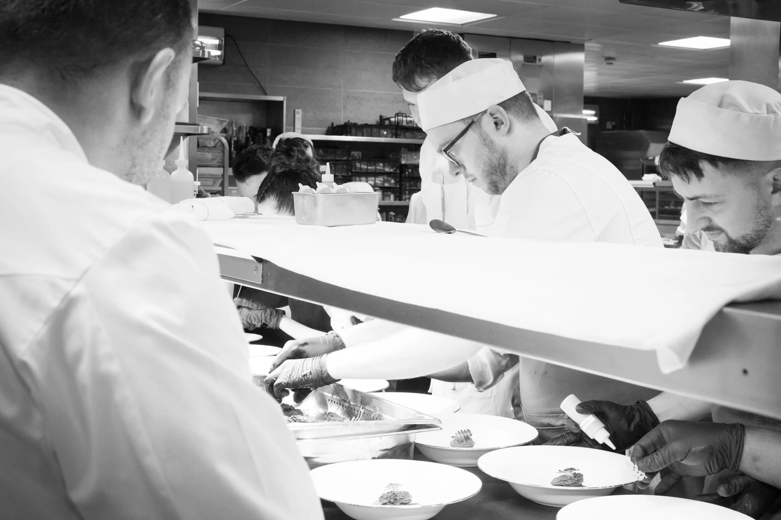 Black and white photo of chefs preparing food in a commercial kitchen, focusing on hands and faces as they work with plates and ingredients.
