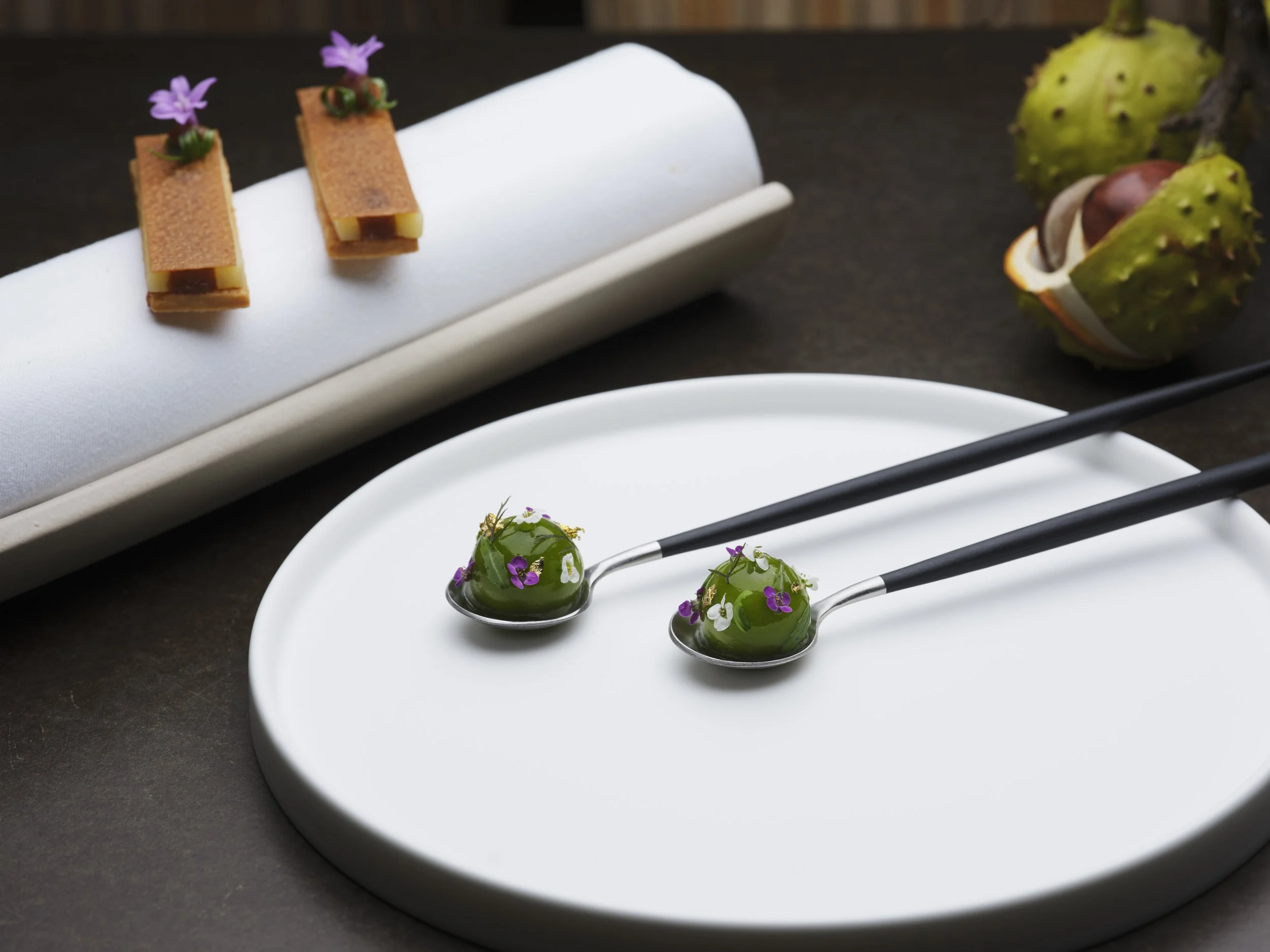 Elegant plated desserts with green jelly topped with edible flowers on a white plate, and small rectangular pastries decorated with purple flowers on a white tray, alongside a spiky green fruit with yellow and purple accents.