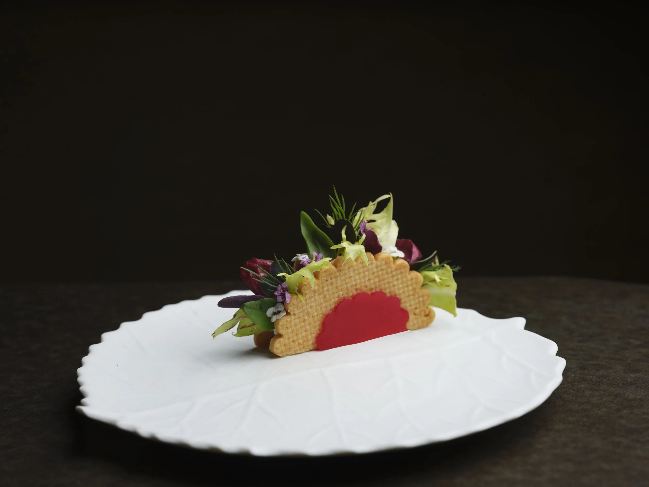 A gourmet dish featuring a decorative arrangement of edible flowers, herbs, a wafer, and a red sauce on a white scalloped plate, with a plain black background.