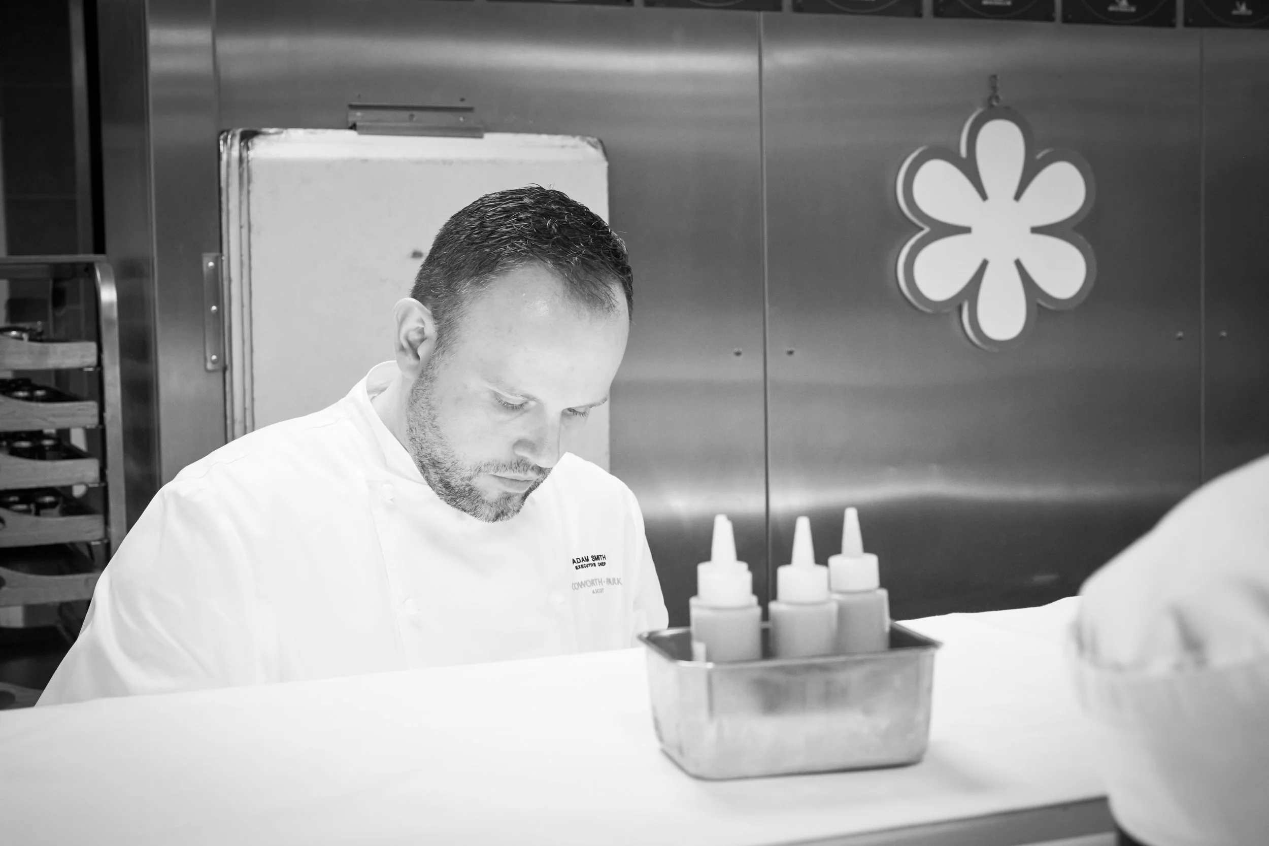 A chef in a white uniform working in a professional kitchen, with a stainless steel wall and bottles on the counter. Behind him is Michelin logo on the wall.