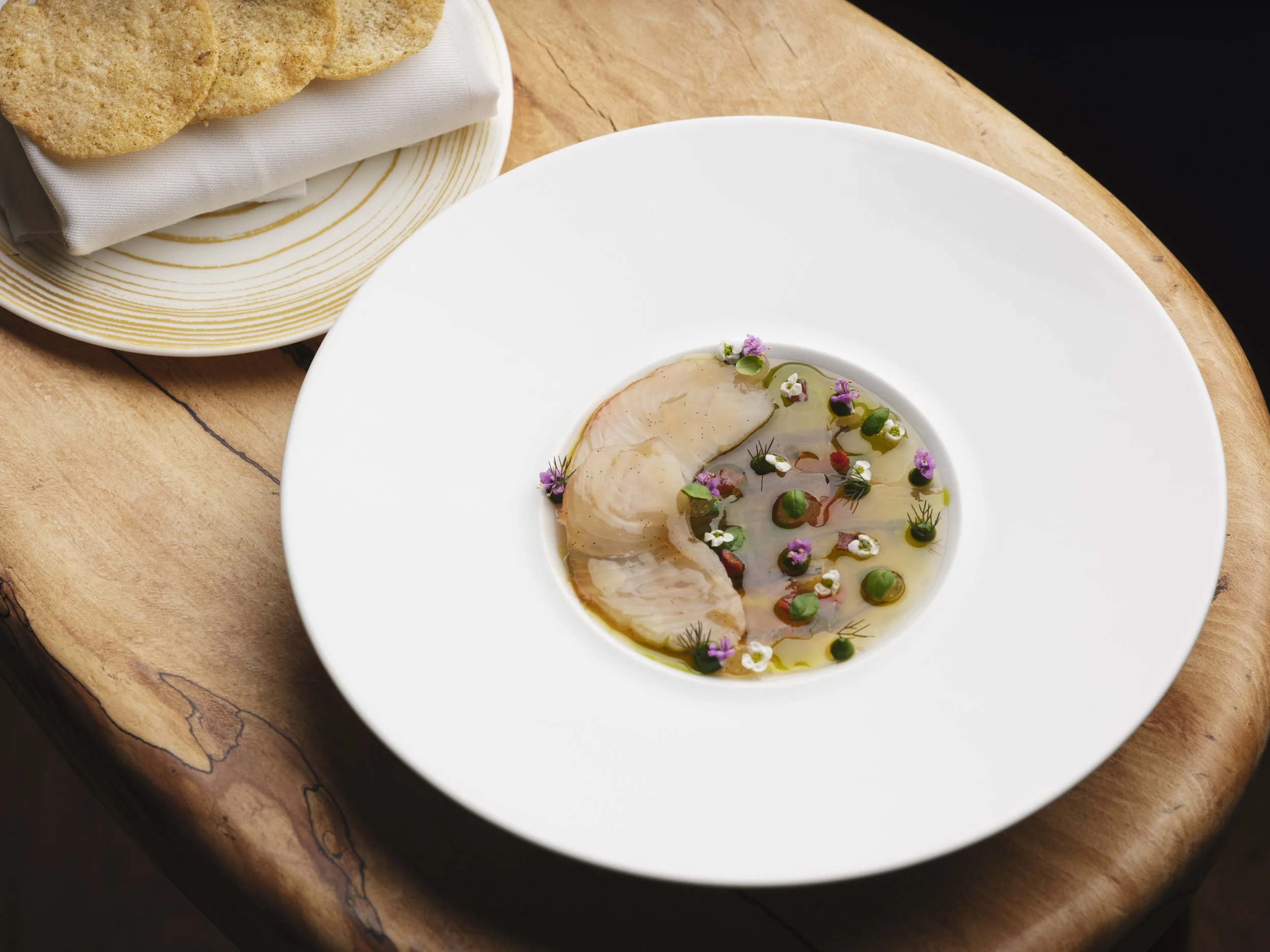 A white plate with sliced fish garnished with herbs, edible flowers, and sauce, placed on a wooden surface, with a plate of flatbread or crackers in the background.