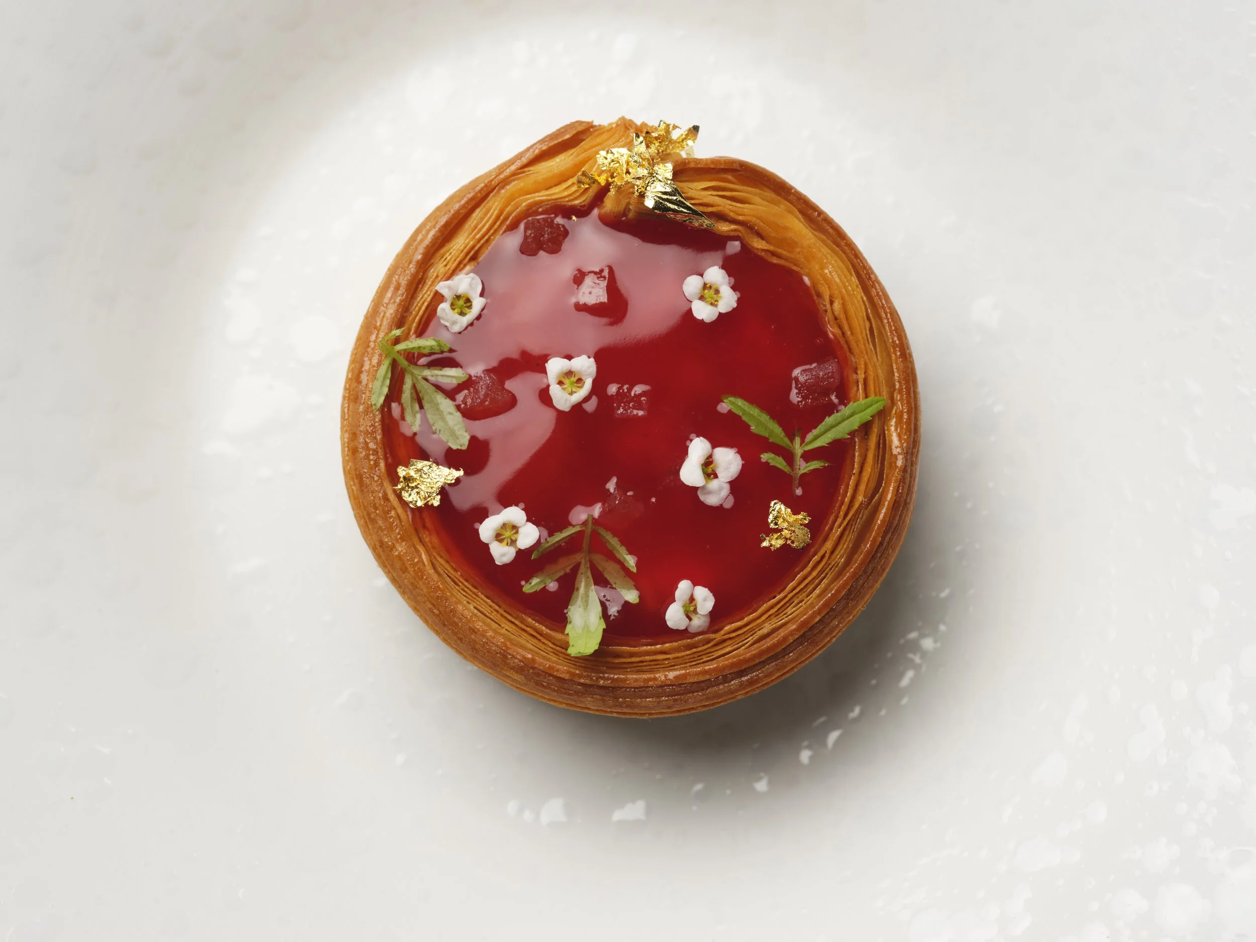 A top-down view of a pastry with strawberr glaze, garnished with small white flowers, green leaves, and edible gold leaf, placed on a white surface.