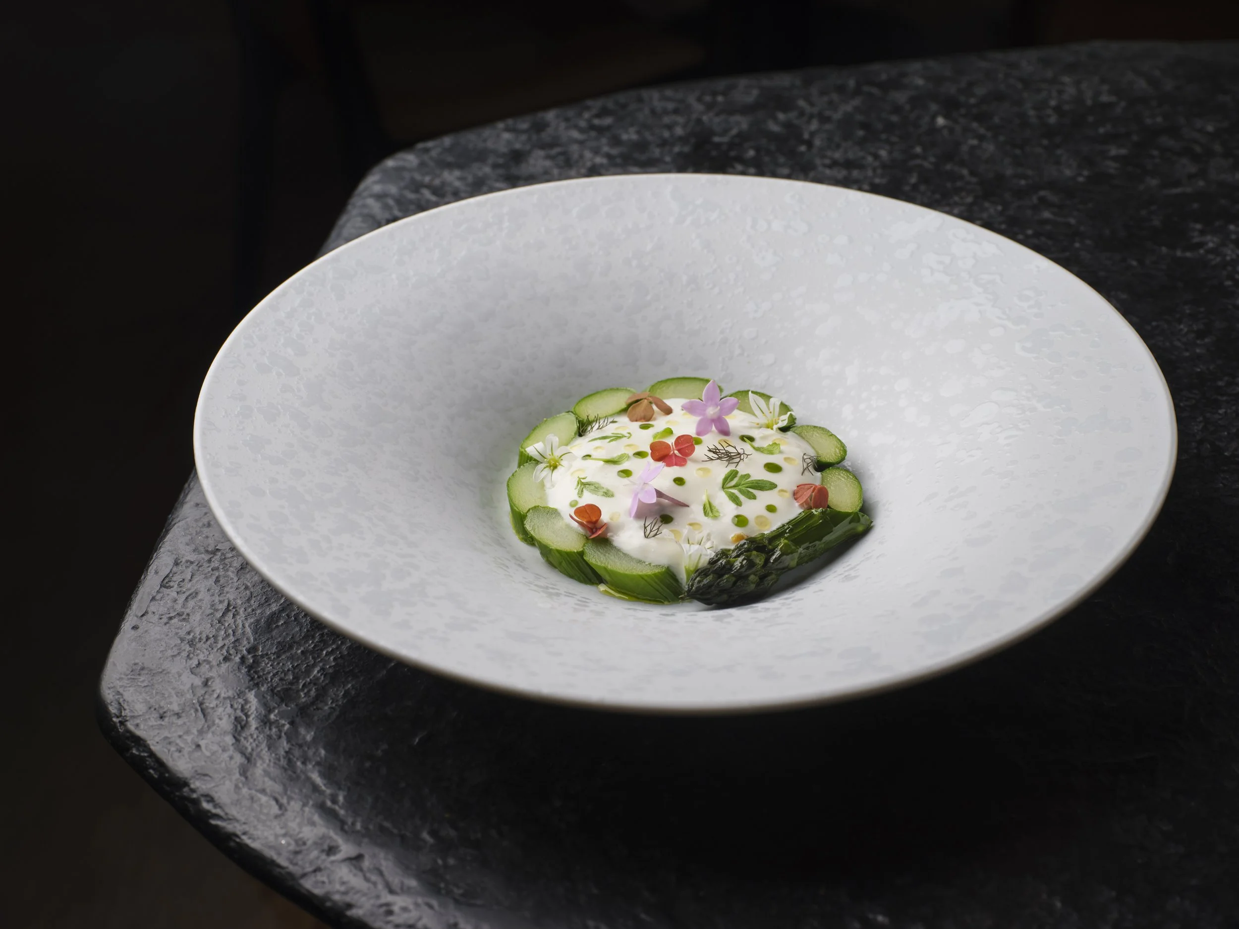 A gourmet dish served on a textured white plate, composed of a circular white base topped with thinly sliced cucumber, edible flowers, green herbs, and small colorful garnishes, positioned on a black granite countertop.
