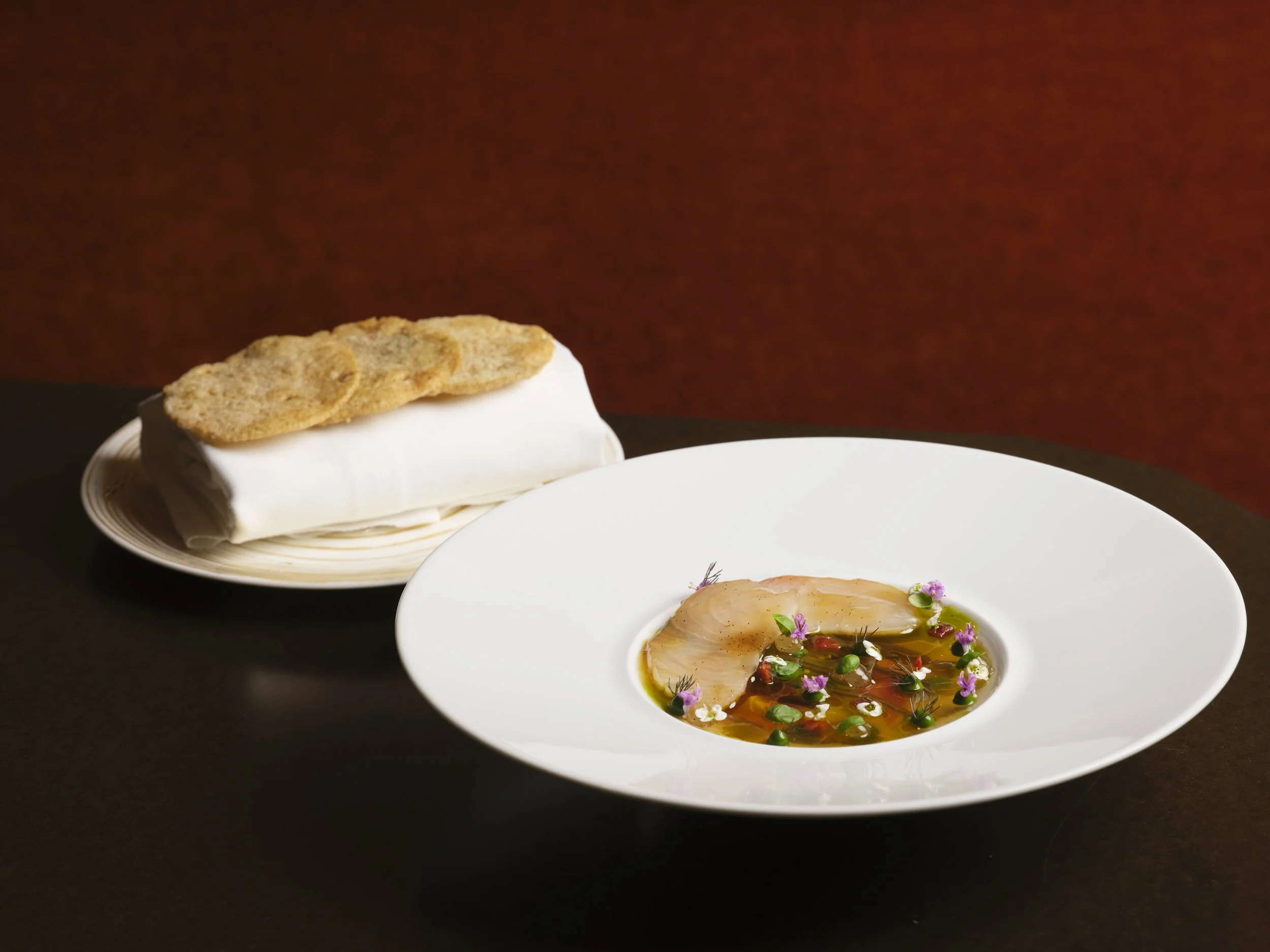 A bowl of sliced raw fish garnished with herbs and edible flowers, served in a light broth, with a plate of crispy bread on the side, set on a dark table with a red background.