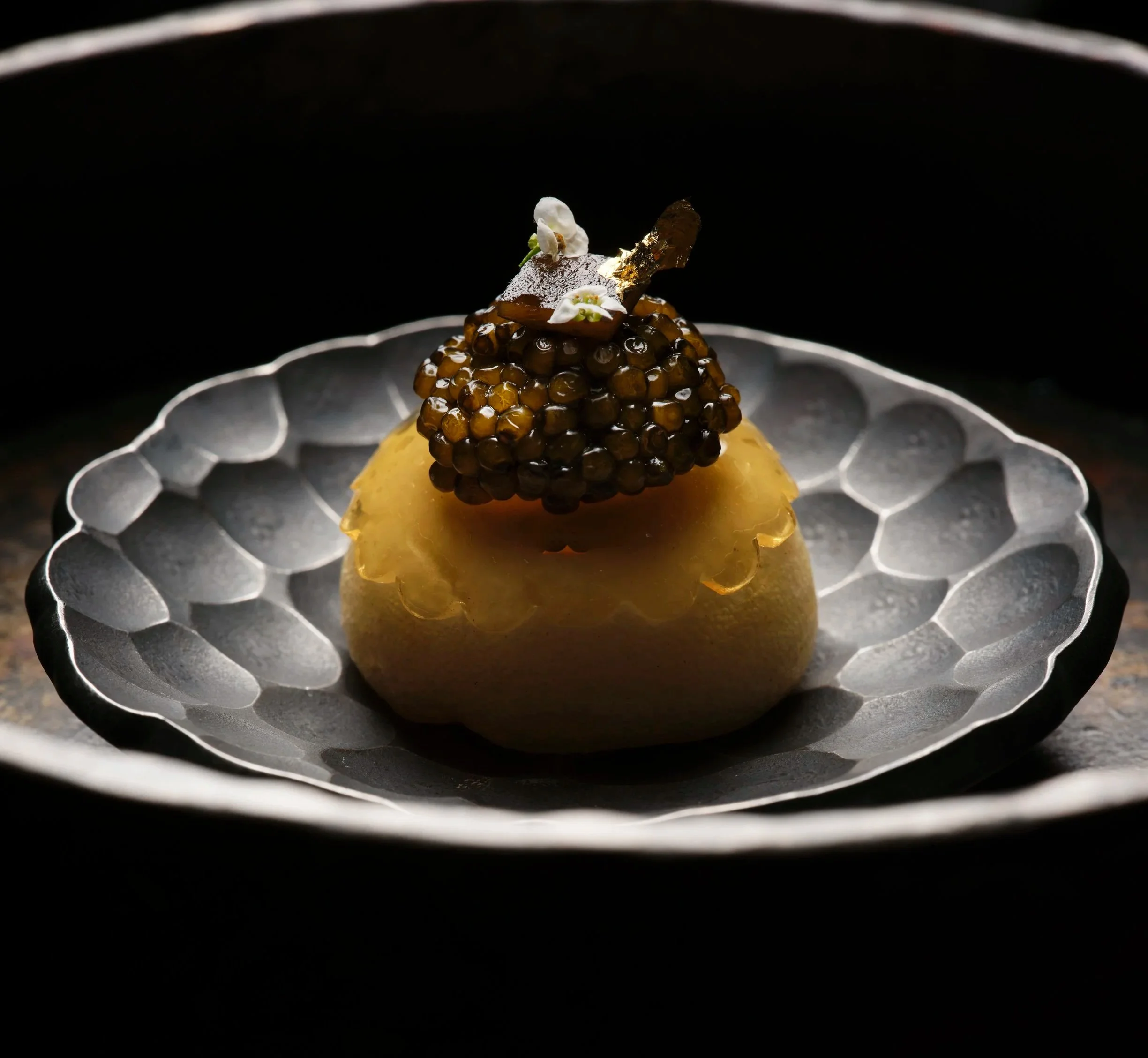 Close-up of a decorative food item featuring black caviar on top of a yellowish base, garnished with a small white flower and a piece of edible gold leaf, presented on a textured silver plate.