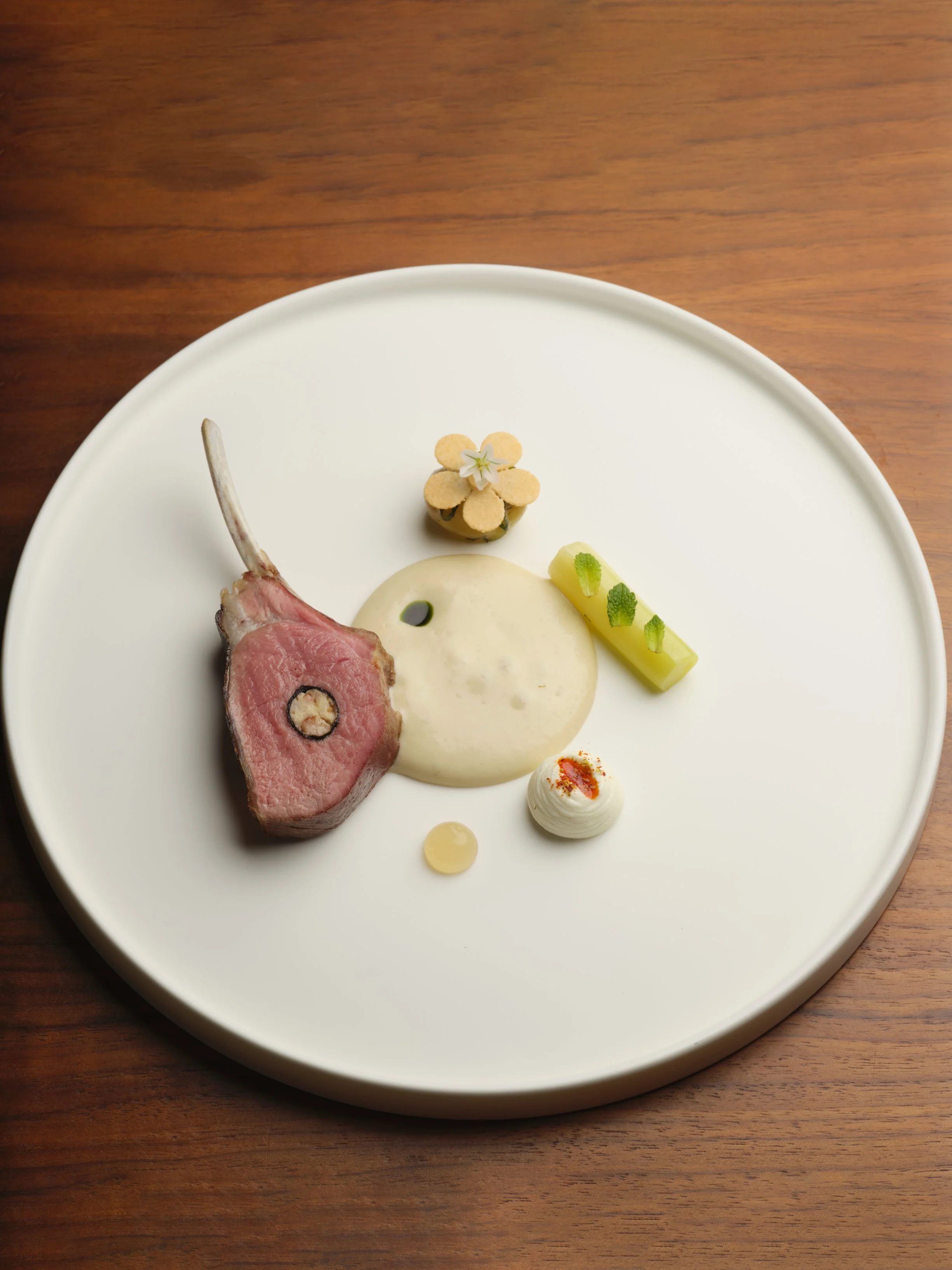 Elegant plated dish with a cut of lamb, a small flower garnish, a scoop of creamy sauce, and assorted small garnishes on a white plate.