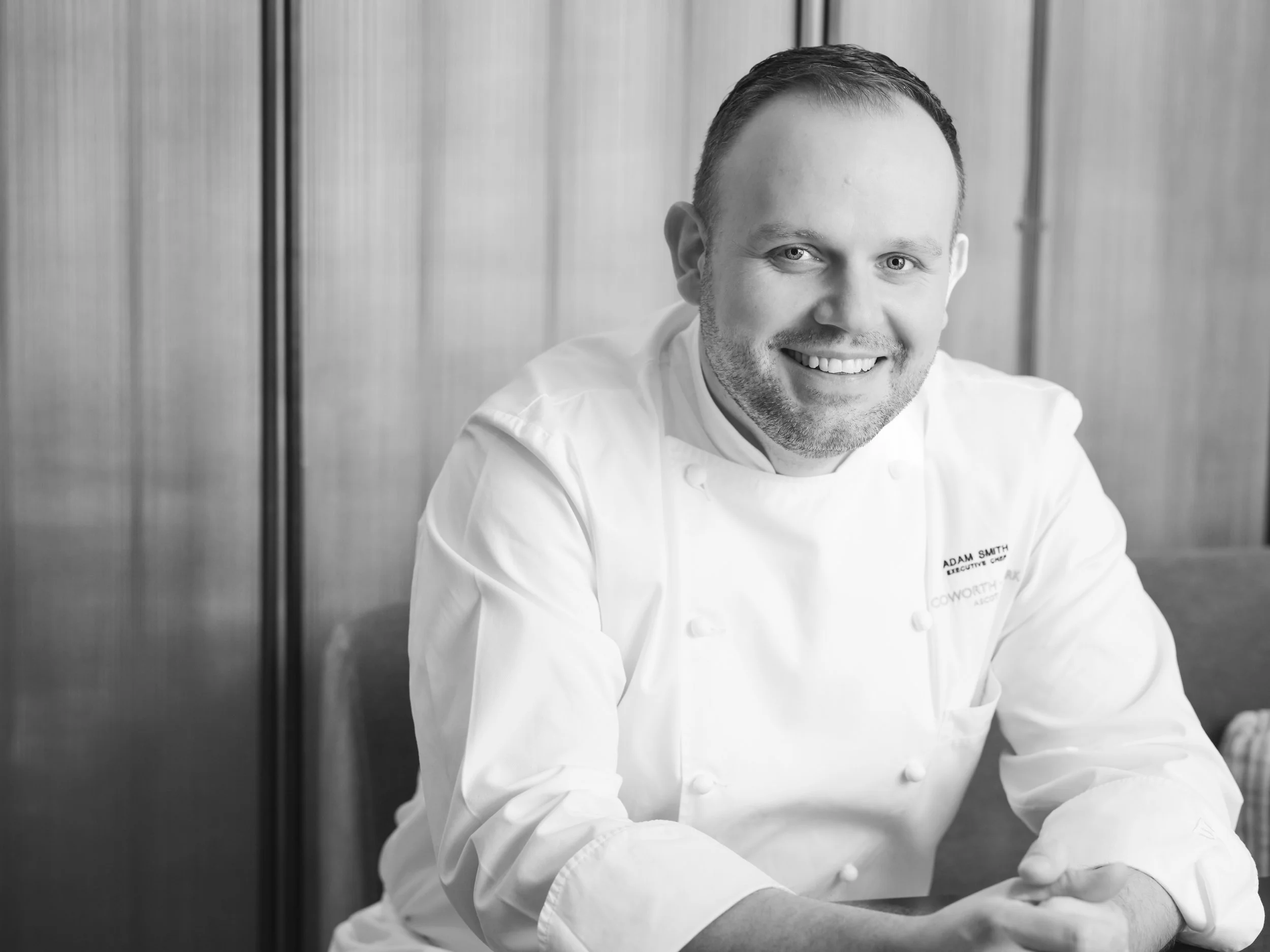 A male chef with short hair and a beard wearing a white chef coat smiling at the camera, in a kitchen setting.