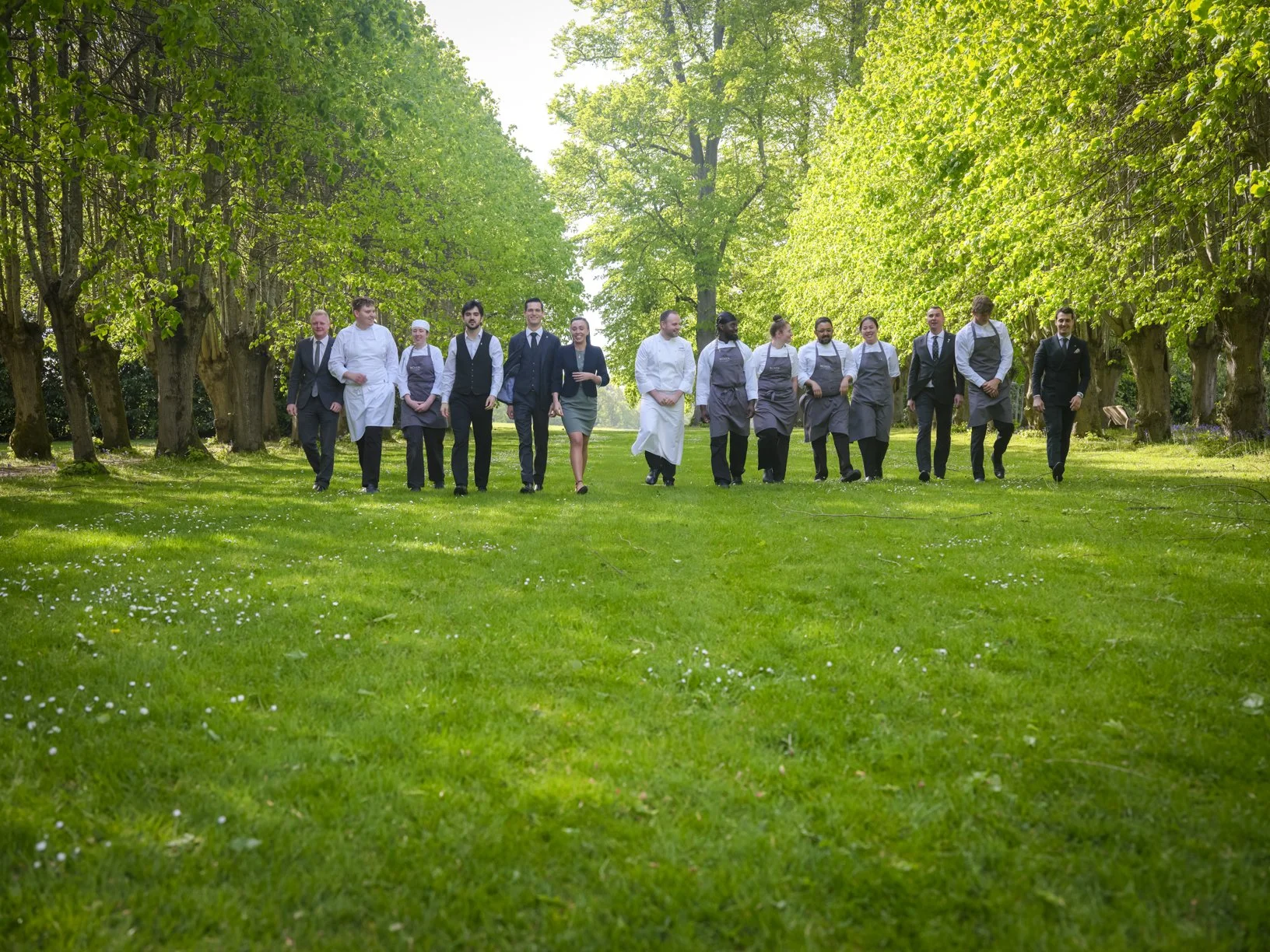 Group of waitstaff and servers walking through a lush green park with tall trees on a sunny day.