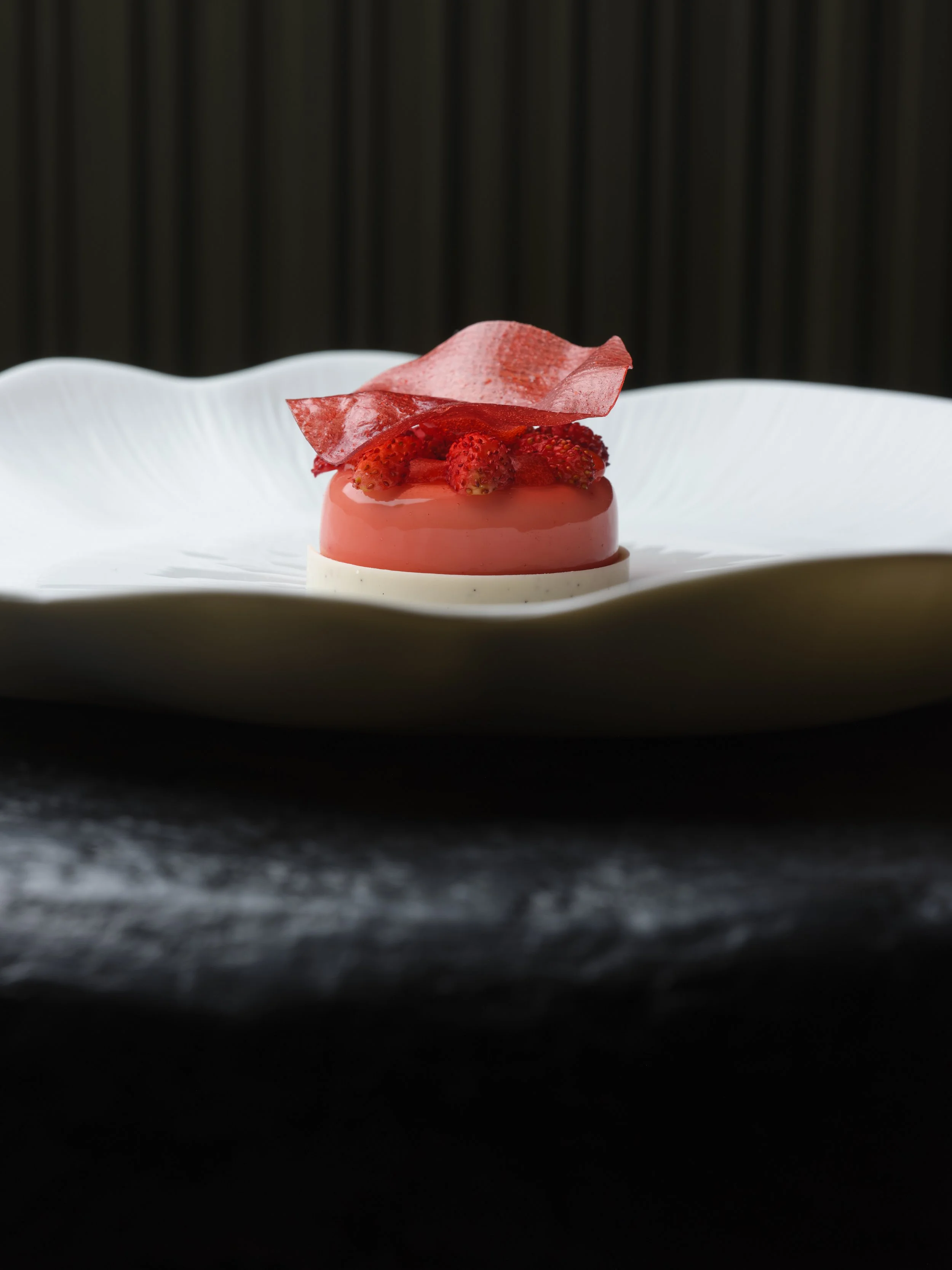 A dessert with a pink glaze topped with fresh strawberries and a pink sugar decoration, served on a white plate with a wavy edge.