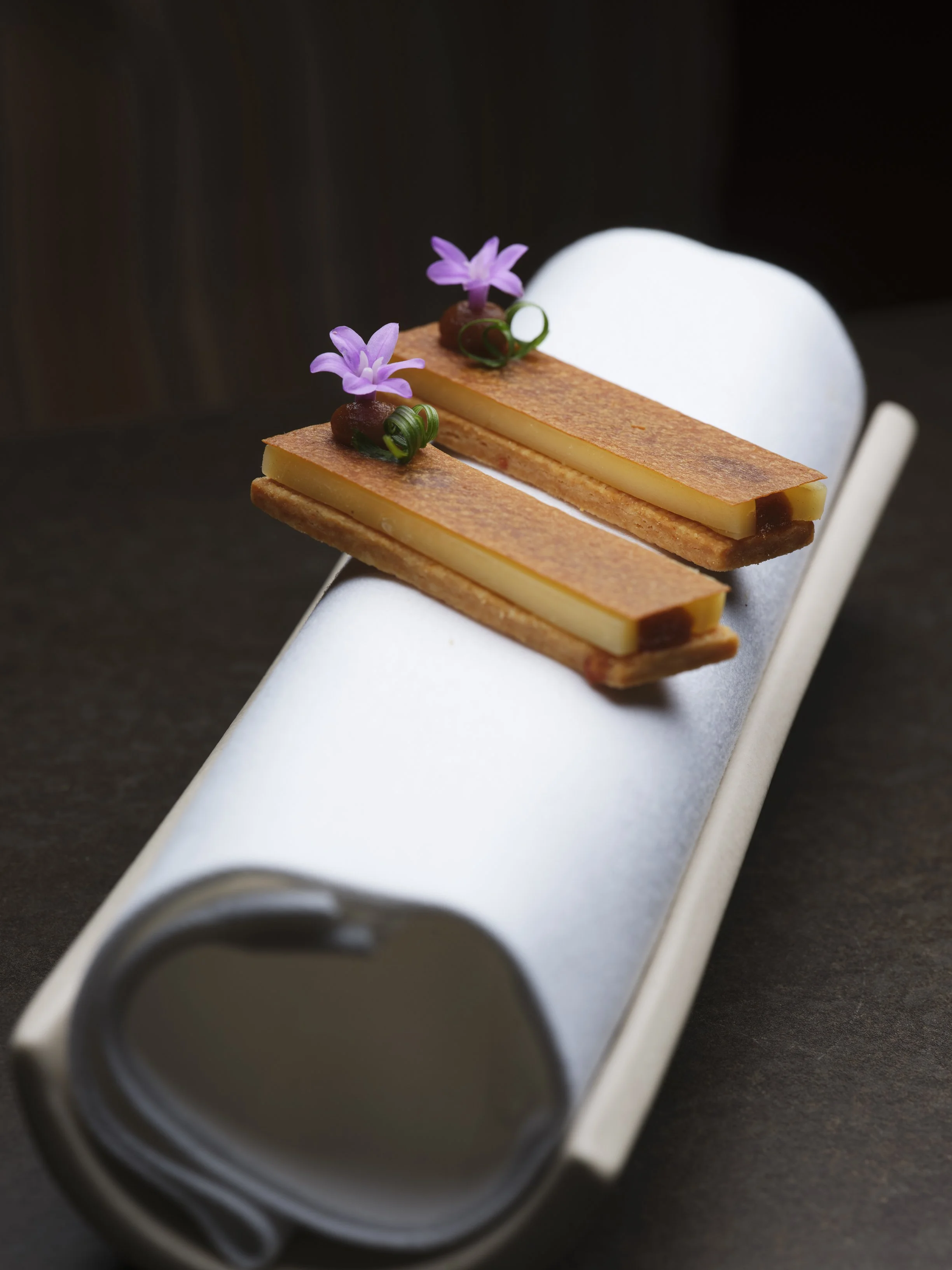 Close-up of a dessert with two rectangular layers of pastry and cream, topped with small decorative purple flowers and green curls, on a silver serving tray.