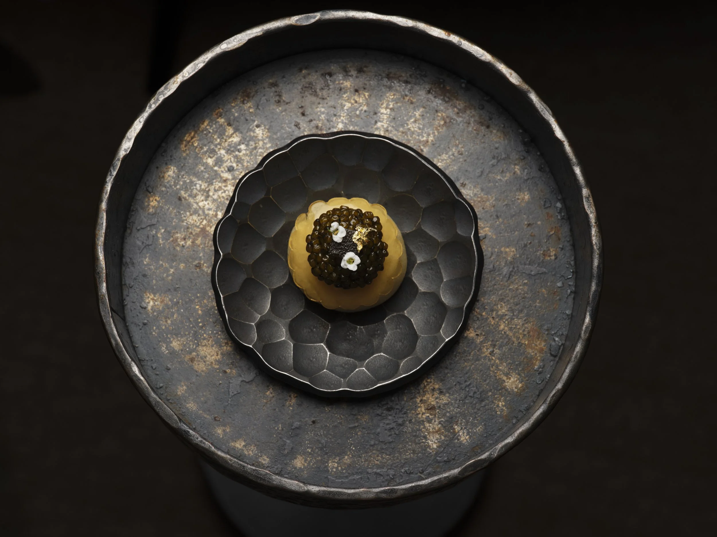 A top-down view of a round, rusted metal surface with a black, honeycomb-patterned bowl in the center. The bowl contains a small, layered canape snack topped with black caviar, edible white flowers, and a gold leaf.