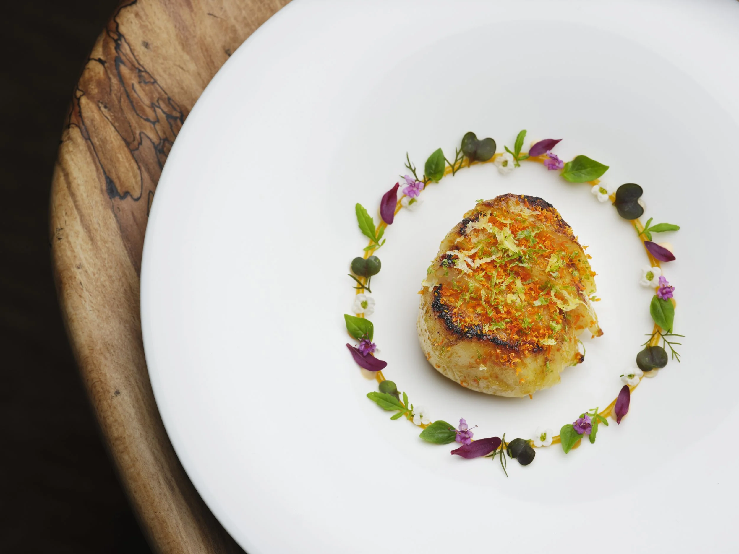 A gourmet dish on a white plate with a wooden board edge visible at the top left. The dish is a seared scallop, surrounded by edible flowers and small green leaves, decorated with a ring of tiny white, pink, purple, and black accents.