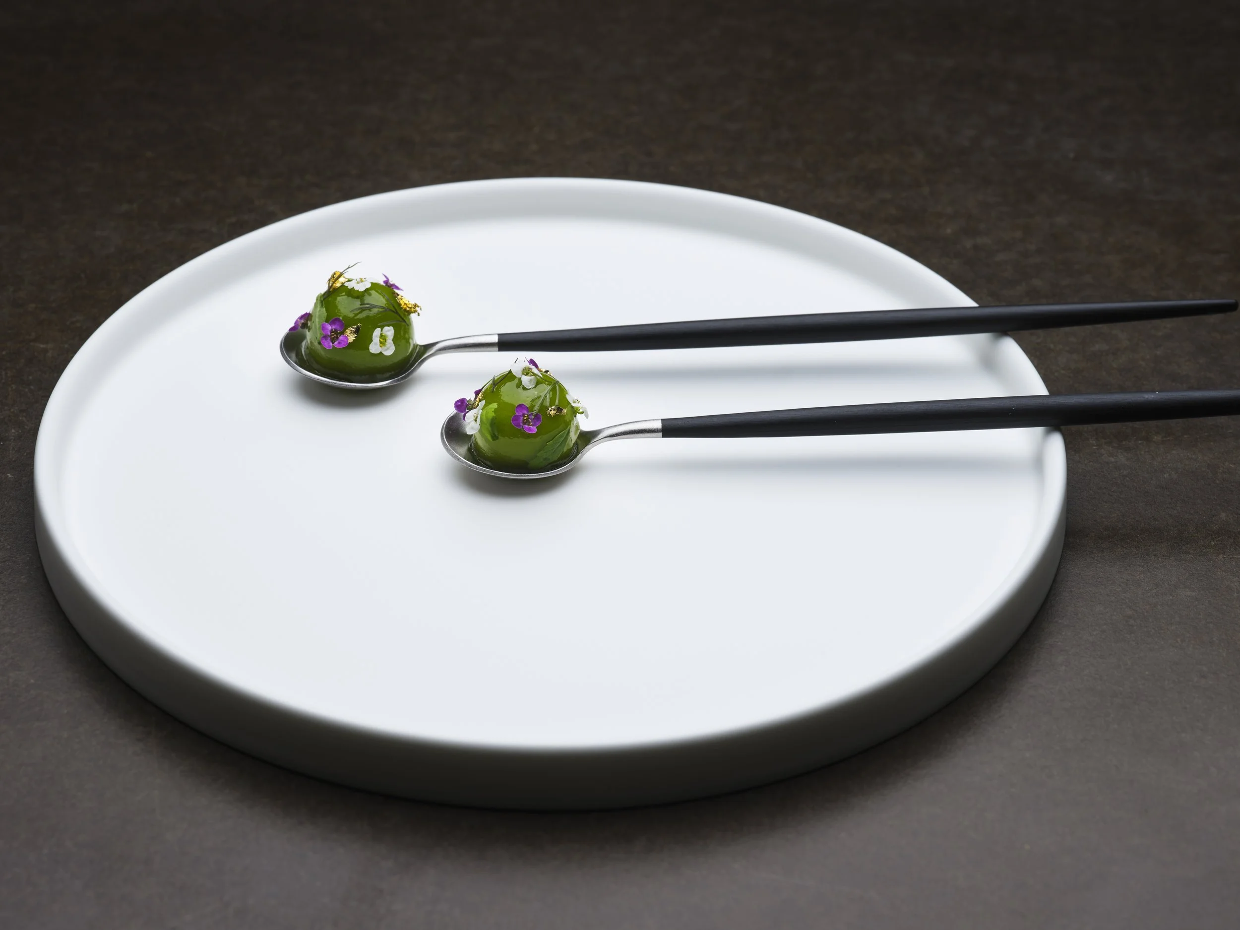 Two white spoons with canape of avocado basil and yuzutopped with small purple and white edible flowers, resting on the edge of a white plate on a dark surface.