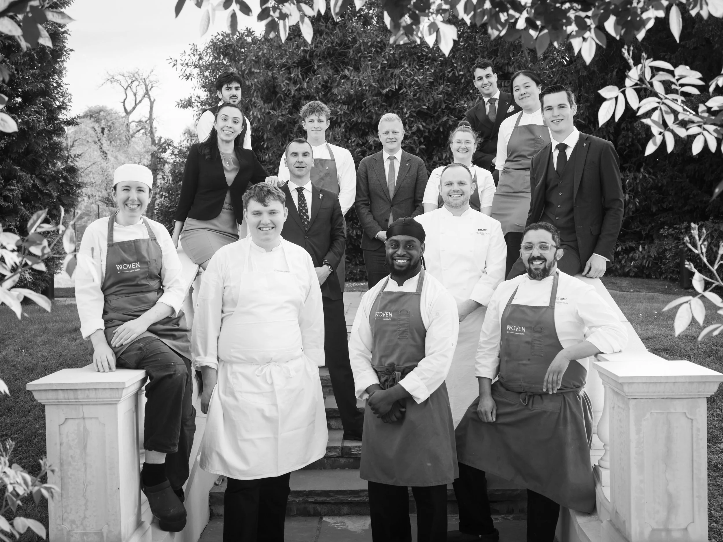 Black and white photo of a diverse group of chefs and restaurant staff posing outdoors on steps, surrounded by trees.