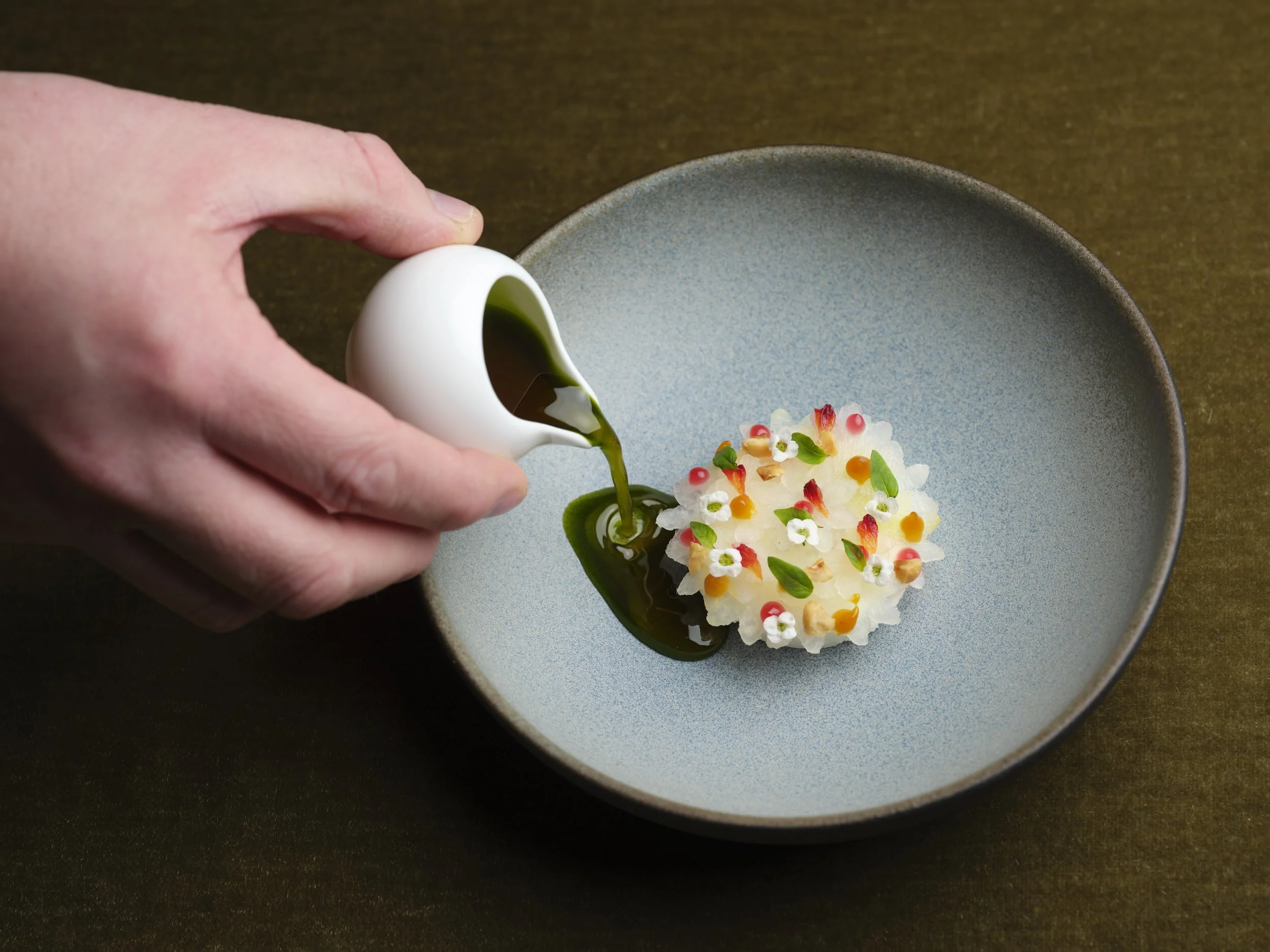 A person is pouring green sauce from a white container onto a kohlrabi dish with flowers and small garnishes, served on a blue-grey plate. It is Lancashire Kohlrabi with kaluga, walnut and wasabi