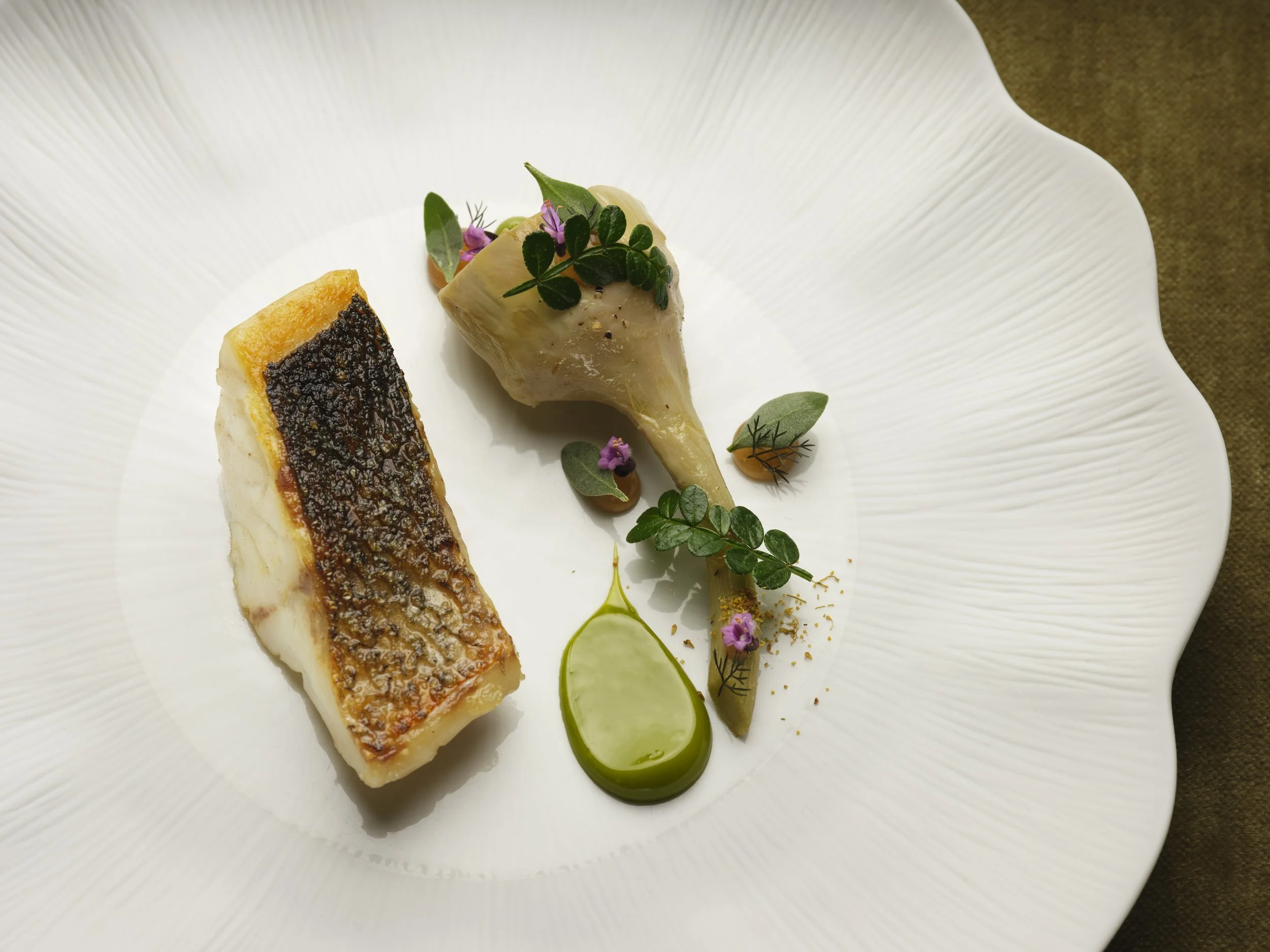 A white plate with two pieces of cooked fish garnished with herbs, edible flowers, and green sauce
