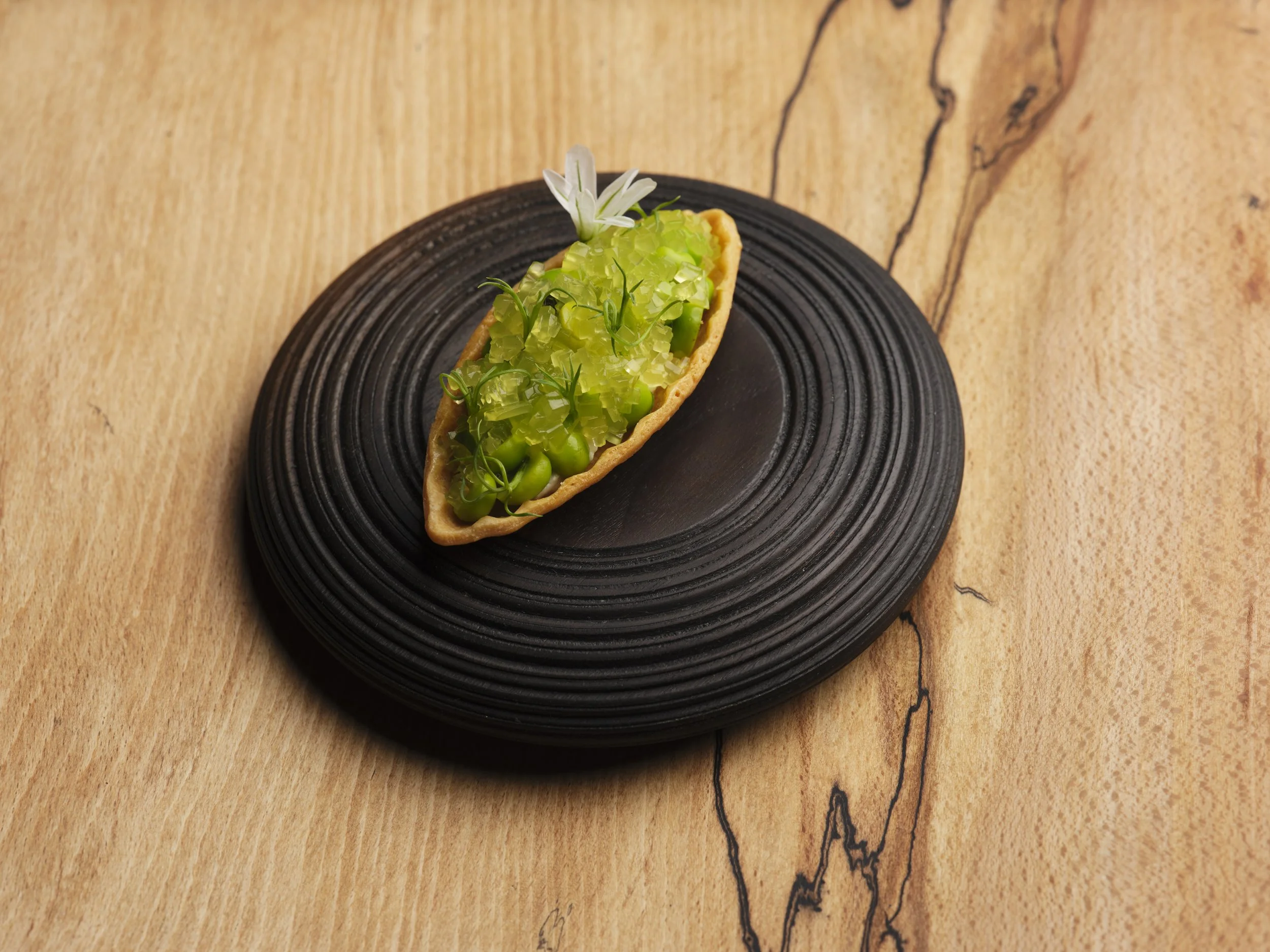A dessert taco with green jelly, green peas, edible flowers, and herbs on a black plate, placed on a wooden surface.
