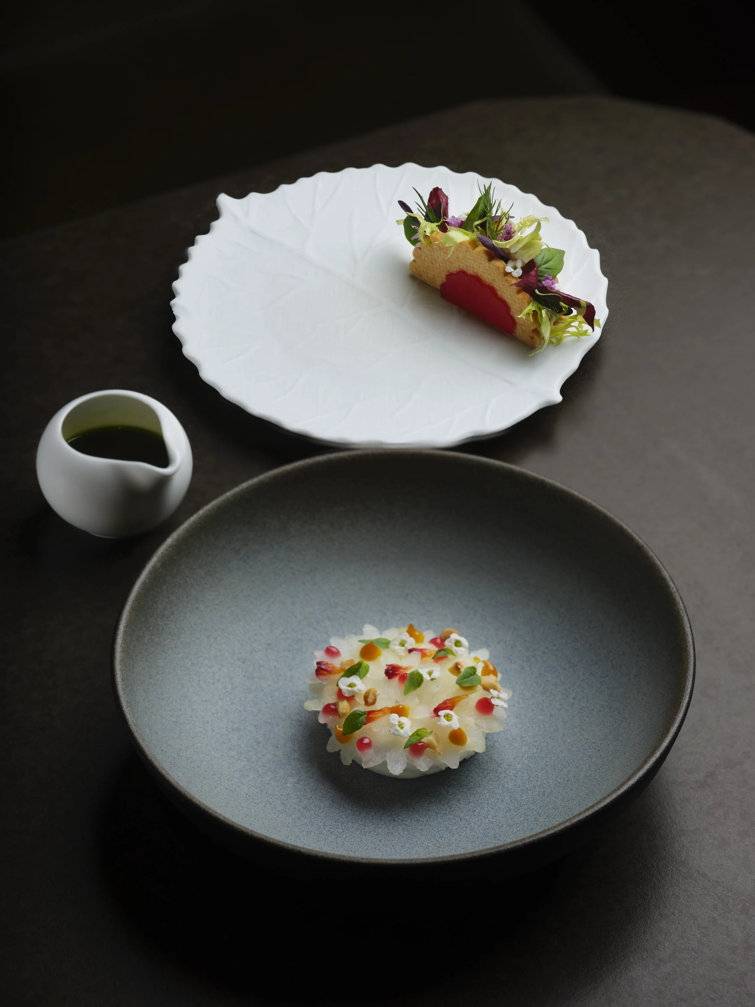 A modern plated dish with a colorful topping on a blue plate, accompanied by a small pitcher of green sauce, on a dark surface.