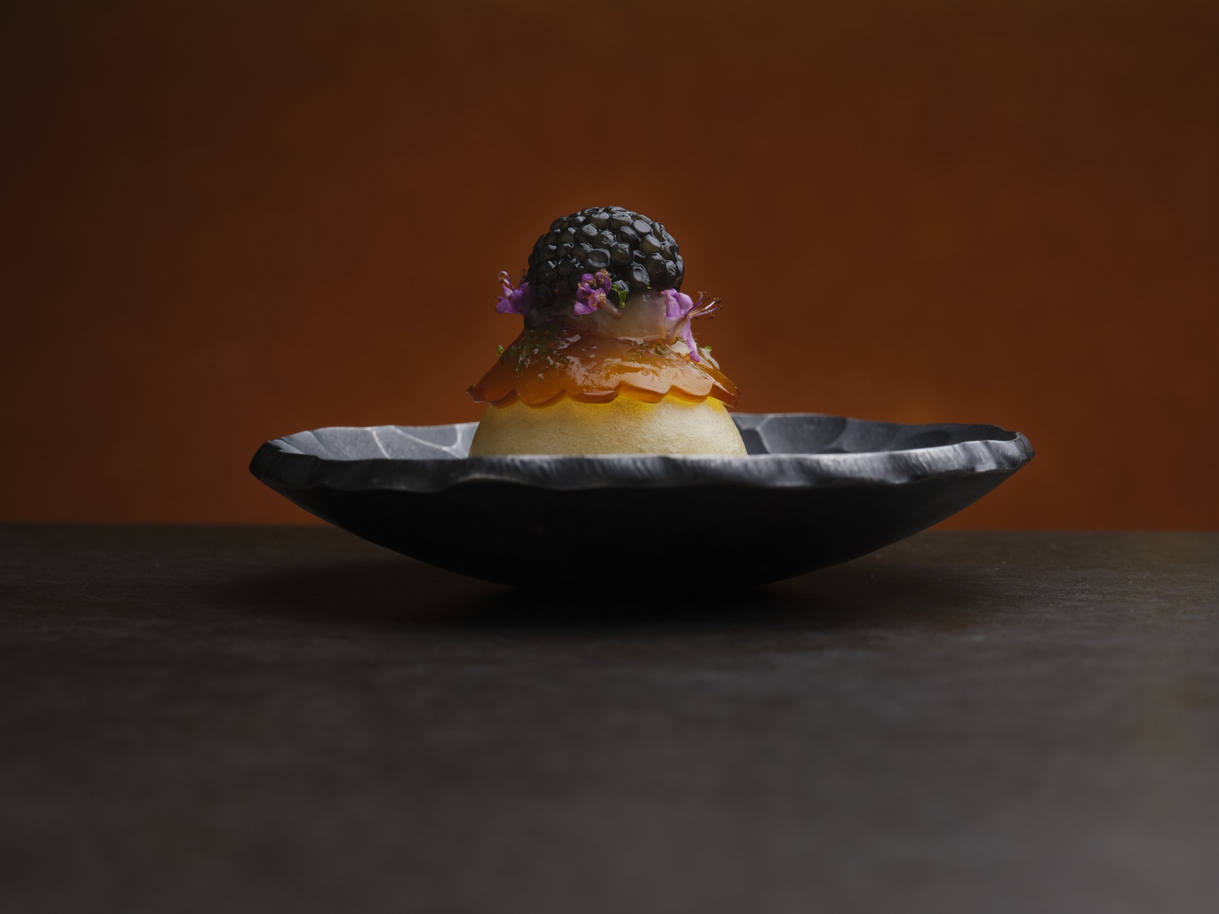 A dessert featuring Dorset crab and Thai green curry topping, garnished with purple flowers, domed with caviar garnish, and set on a black plate with a textured rim. The background is a warm, solid brown colour.