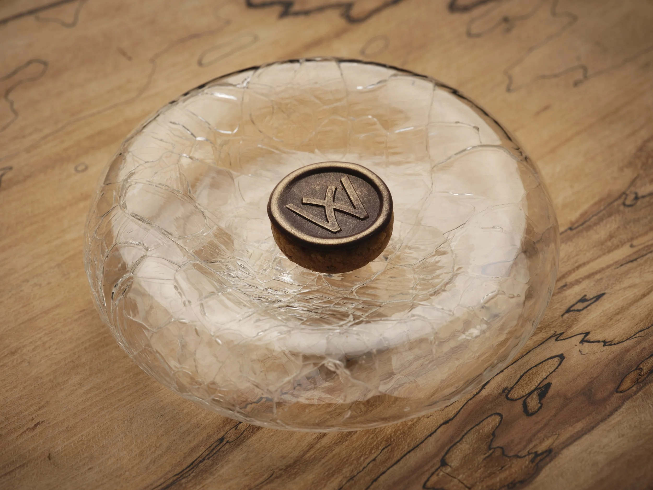 Close-up of a chocolate petit four on a glass container with cracked ice on a wooden surface.