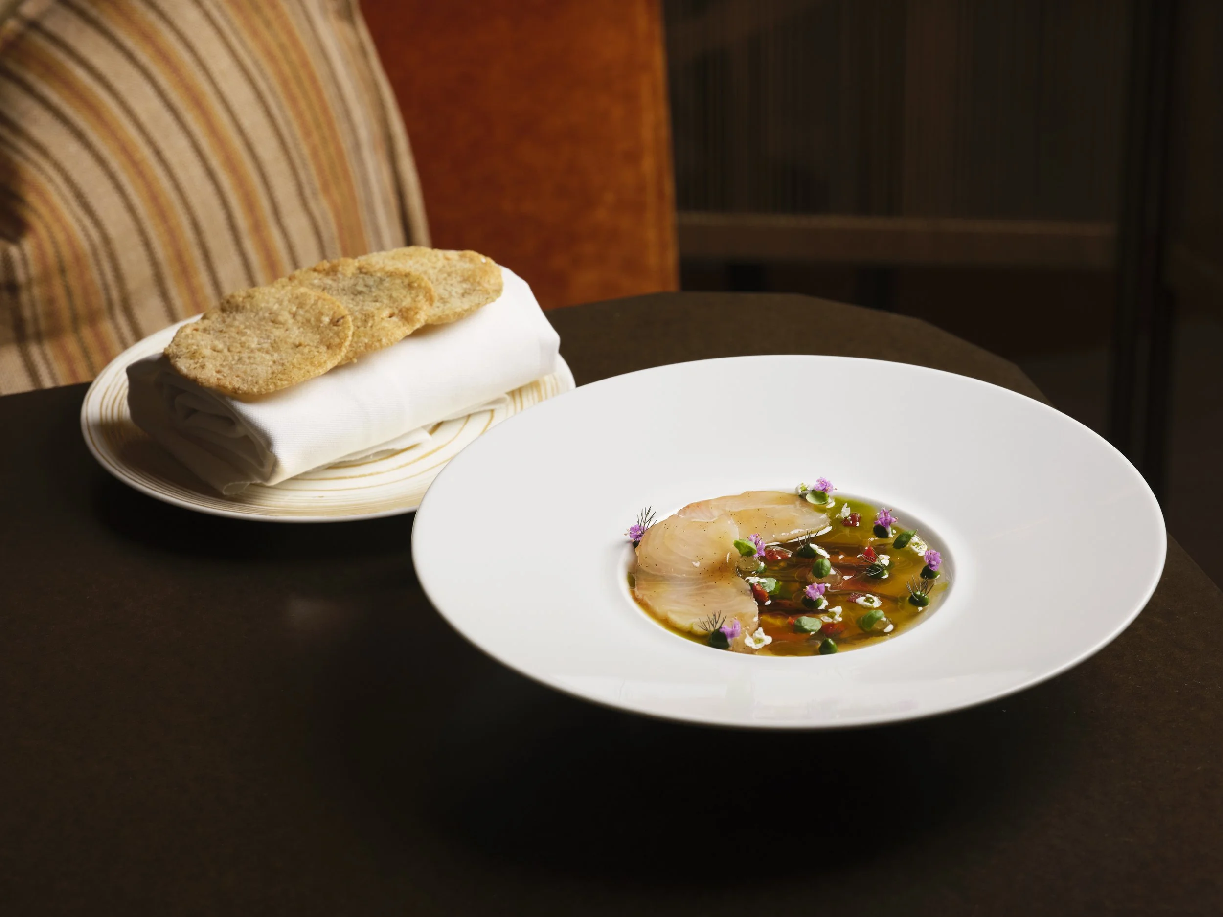 A white plate contains raw fish in a broth garnished with herbs and flowers, next to a plate of sliced bread on a white napkin.