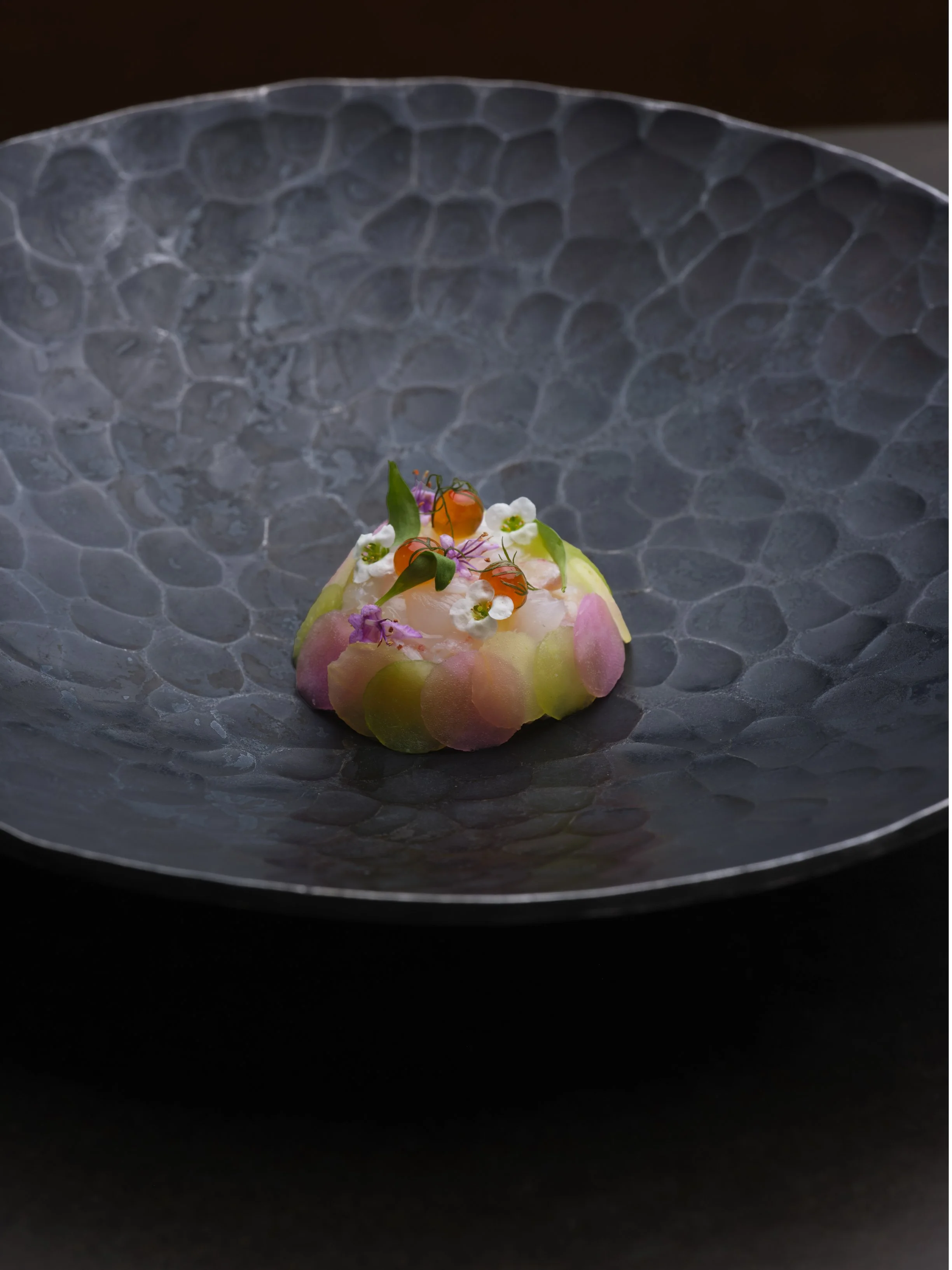 A dessert topped with edible flowers, small berries, and colorful garnishes on a textured black plate.