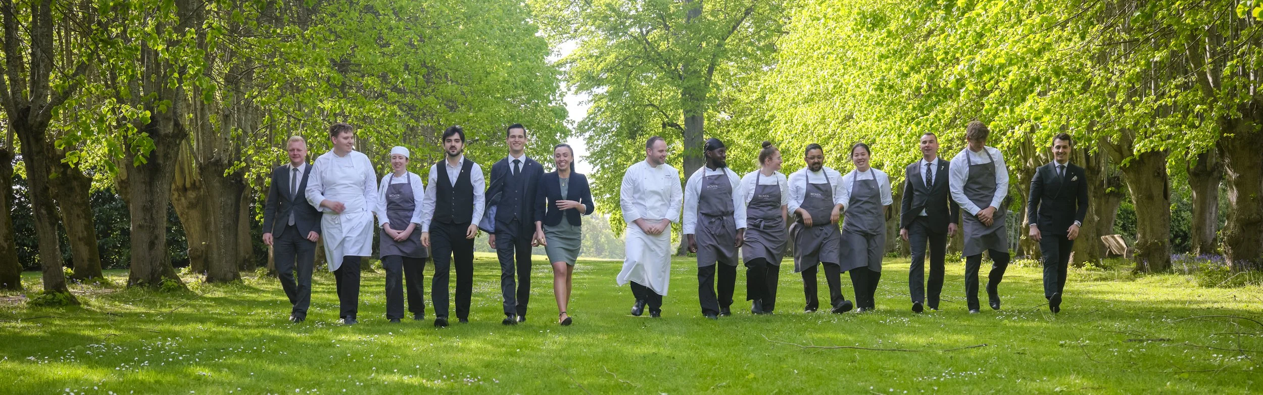 A group of diverse people dressed in professional uniforms and suits walking outdoors through a green, tree-lined park on a sunny day.