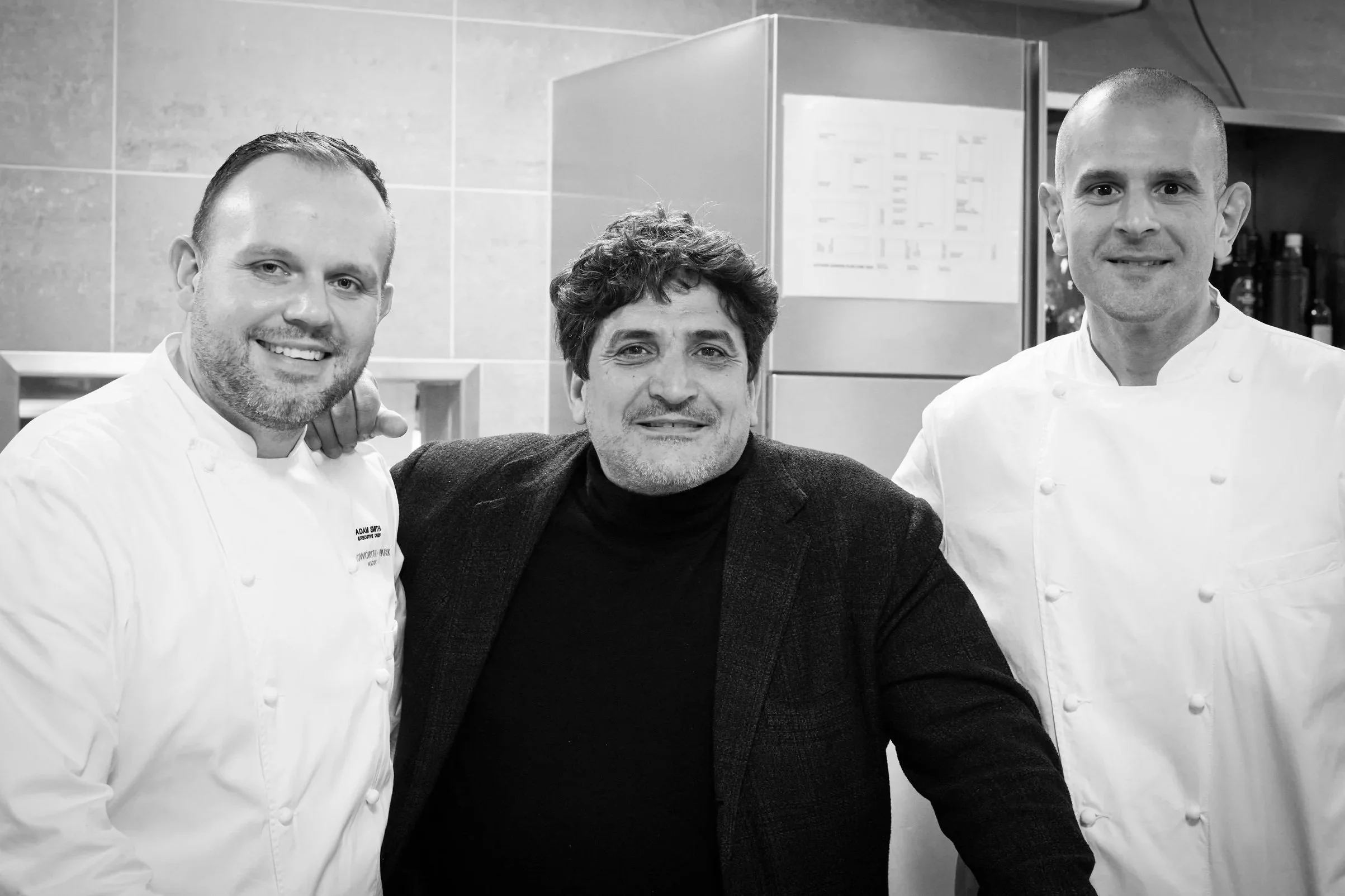 Three men posing together in a kitchen, two are dressed as chefs in white uniforms and one is dressed in a dark blazer. All are smiling and facing the camera.