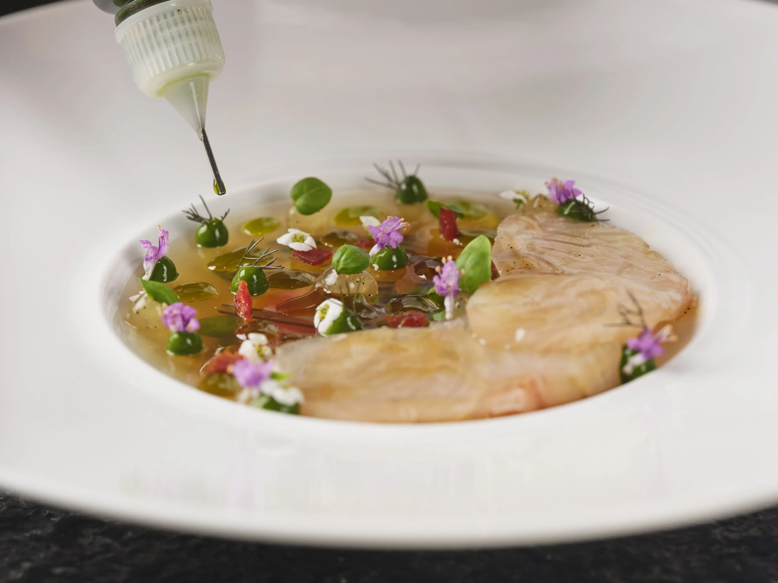 Close-up of a white plate with raw salmon, garnished with green herbs, edible flowers, and drops of sauce. An eyedropper with a green liquid is positioned above the plate, suggesting the addition of flavor or seasoning.