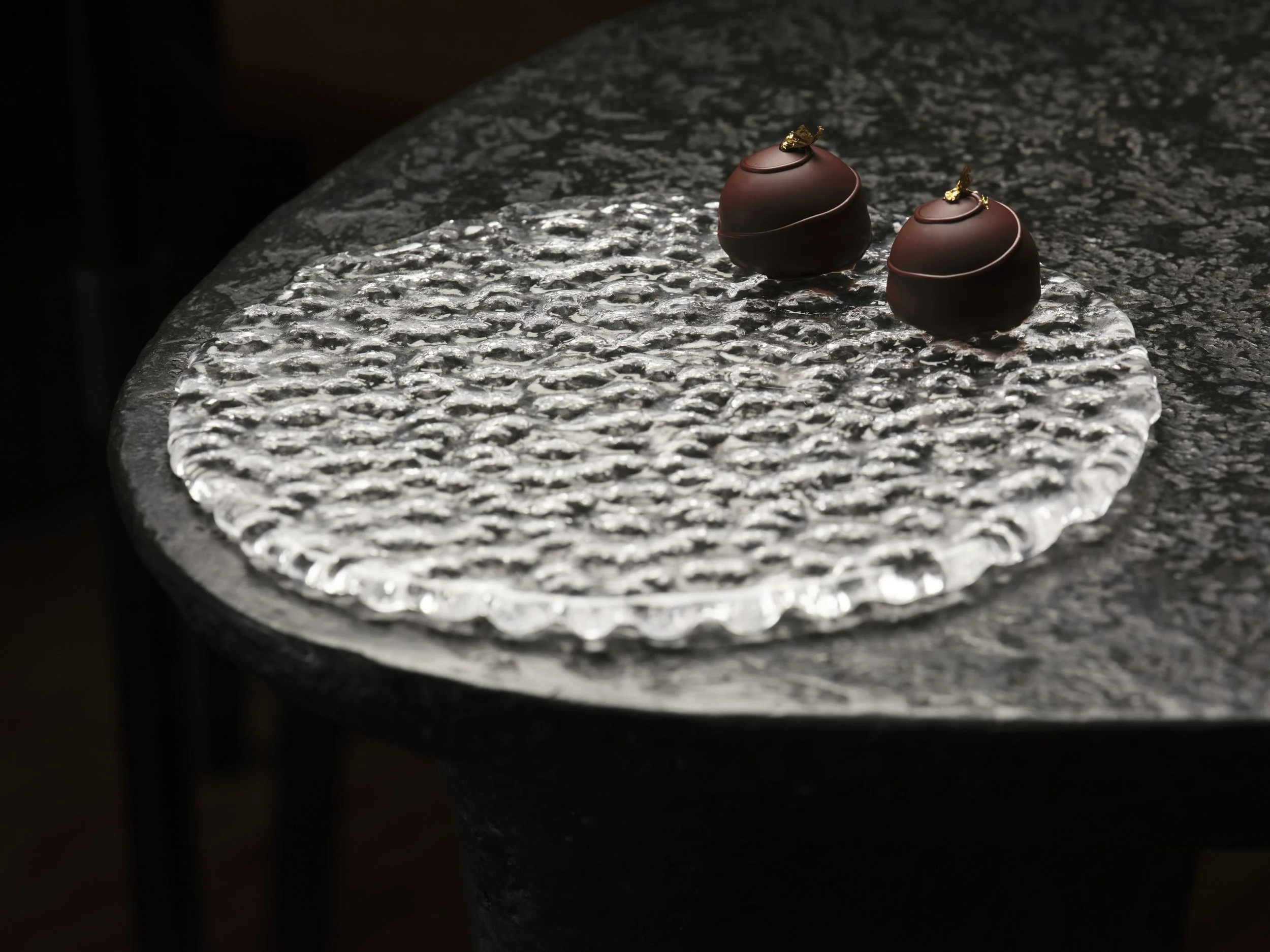 Two chocolate truffles with gold leaf on top resting on a textured metallic surface.