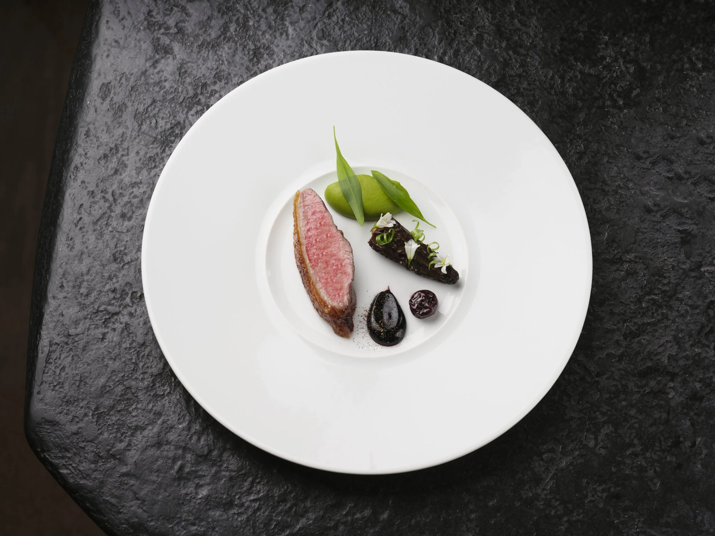 A white round plate with a serving of duck, green vegetables, a dark sauce, and small garnishes, set on a black textured surface.